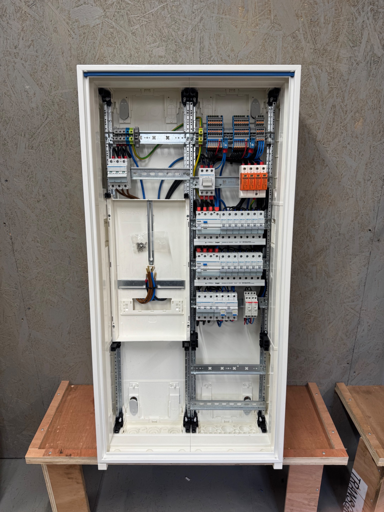 Rozdzielnica elektryczna w białej obudowie, widoczne zabezpieczenia, przewody i szyny montażowe na tle płyty OSB.