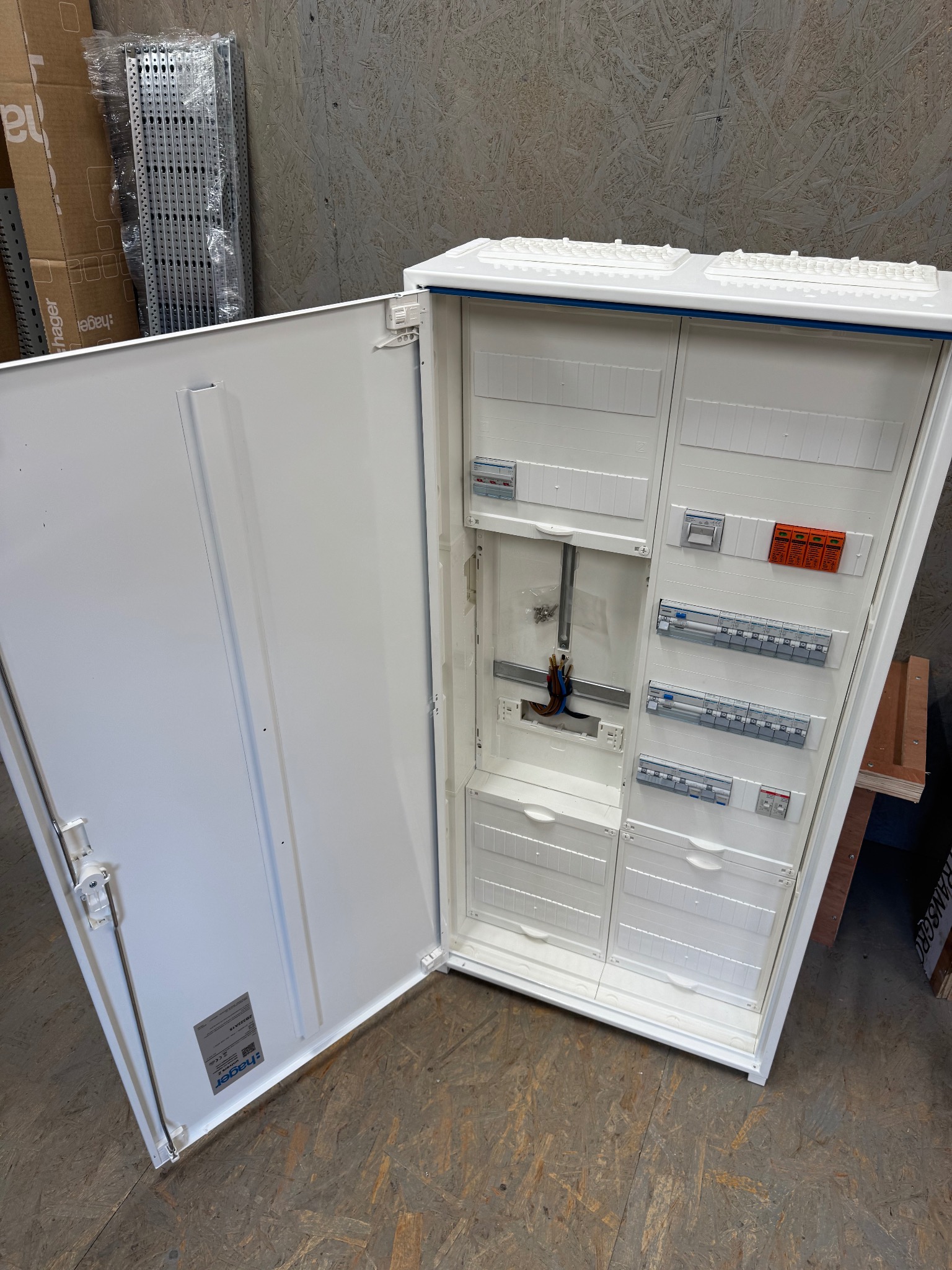 Otwarta, biała skrzynka elektryczna Hager z widocznymi bezpiecznikami i przewodami, na tle drewnianej płyty. Widoczny osprzęt i elementy instalacji.