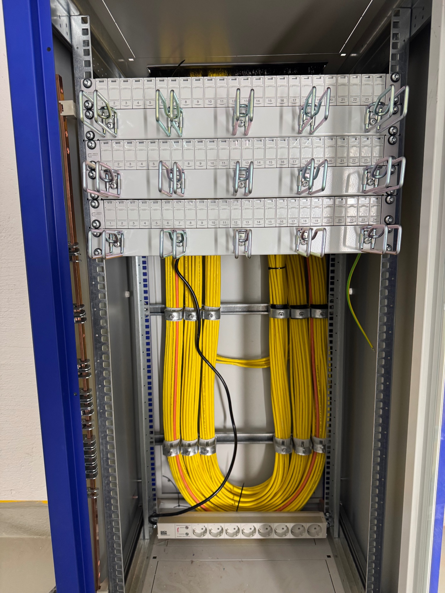Wnętrze szafy rack z uporządkowanymi kablami w żółtej osłonie, listwą zasilającą i panelami krosowniczymi z numeracją, widok z bliska.