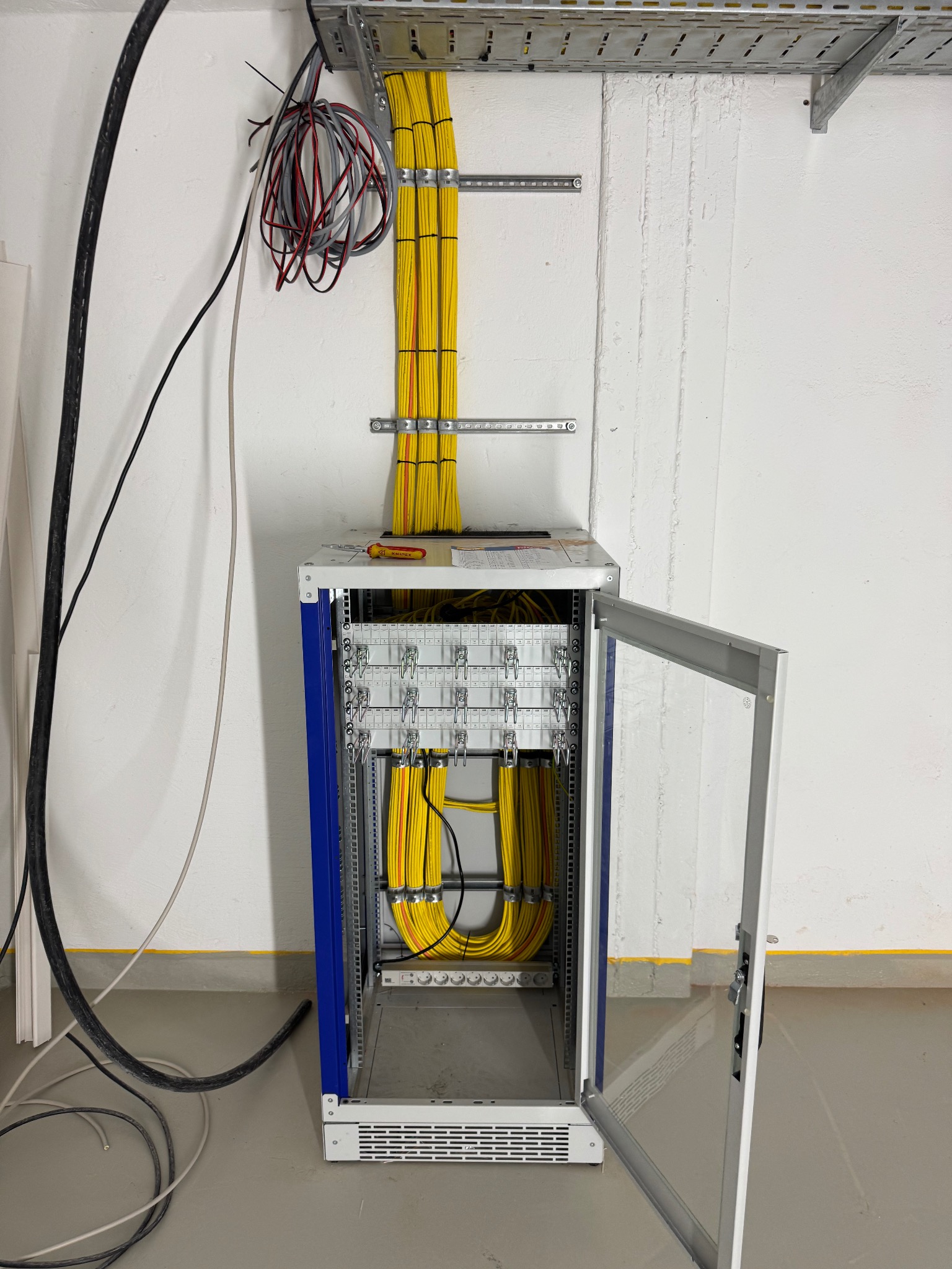 Szafa rack z uporządkowanymi, żółtymi kablami wewnątrz, widoczne narzędzia na górze szafy, instalacja kablowa na ścianie. Otwarta przeszklona obudowa.