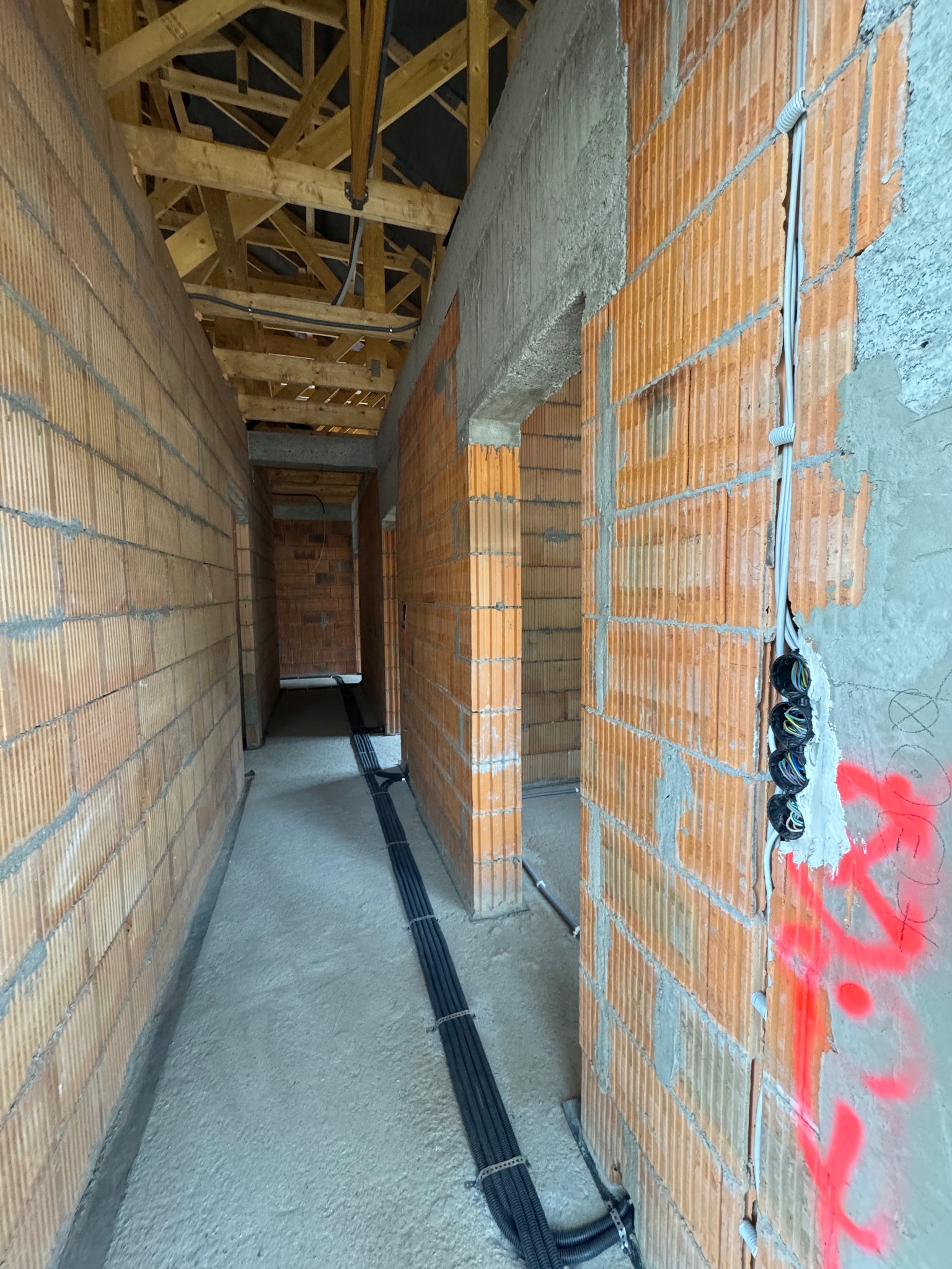 Wnętrze w trakcie budowy: widok korytarza z instalacją elektryczną w bruzdach ścian z cegły, widoczne kable w peszlach i puszki elektryczne, surowy beton i drewniana konstrukcja dachu.