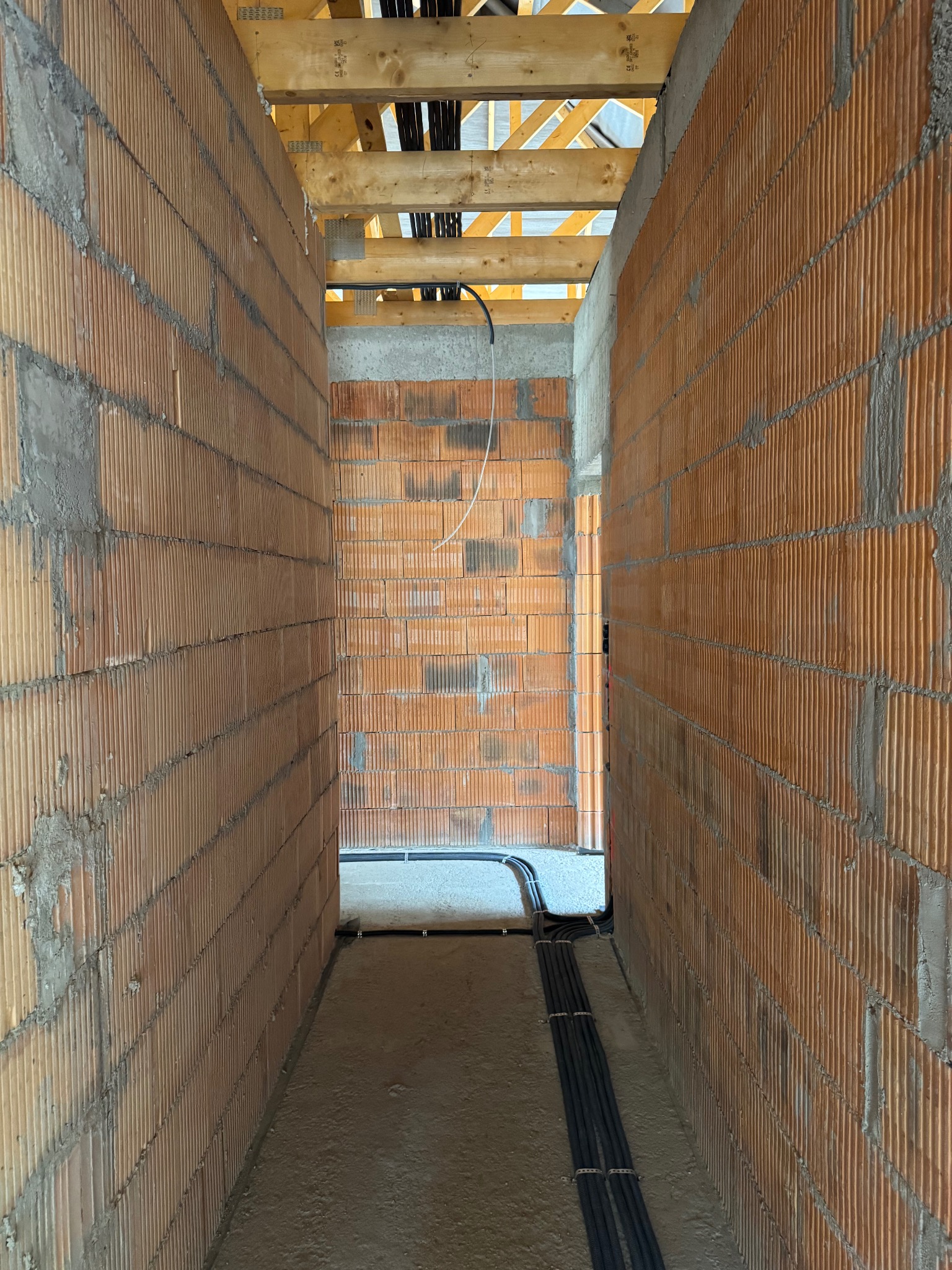 Wnętrze w trakcie budowy z widocznymi przewodami elektrycznymi ułożonymi wzdłuż podłogi i ścian z cegły. Widoczna konstrukcja drewniana stropu.