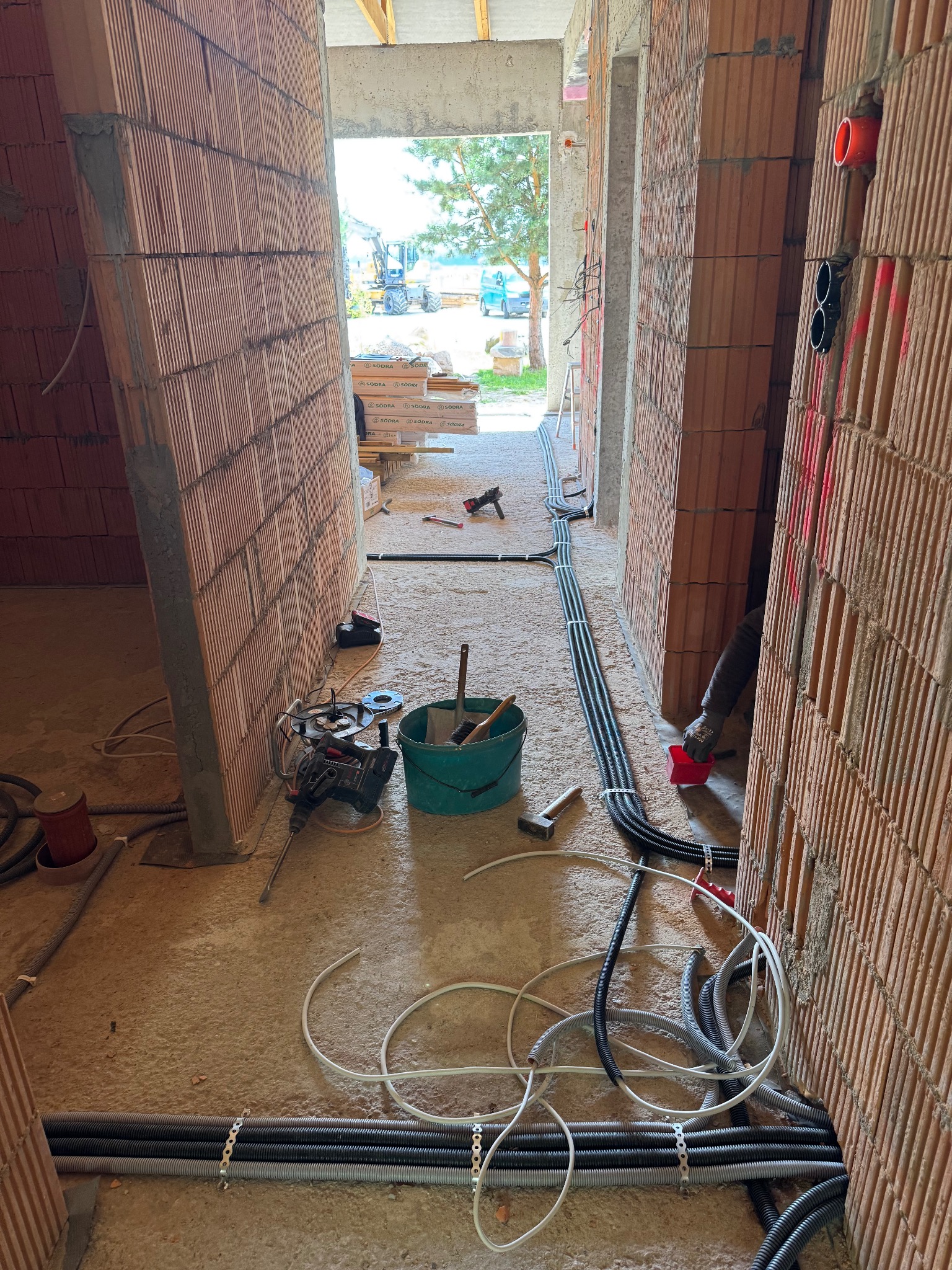Wnętrze w trakcie budowy: ściany z cegły, narzędzia (wiertarka, młotek), kable prowadzone wzdłuż ścian i podłogi, widok na zewnątrz przez otwór w ścianie.