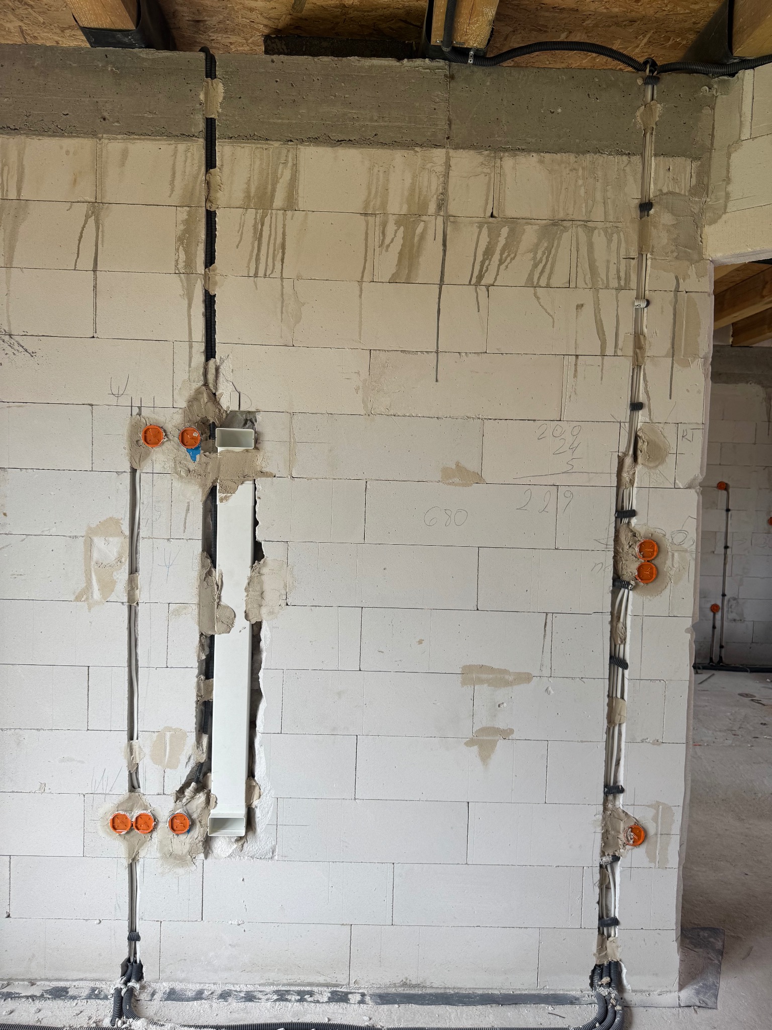 Biała ściana z betonu komórkowego z wyżłobieniami na kable elektryczne i pomarańczowymi puszkami instalacyjnymi. Widoczne przewody w peszlach i rurkach PCV.
