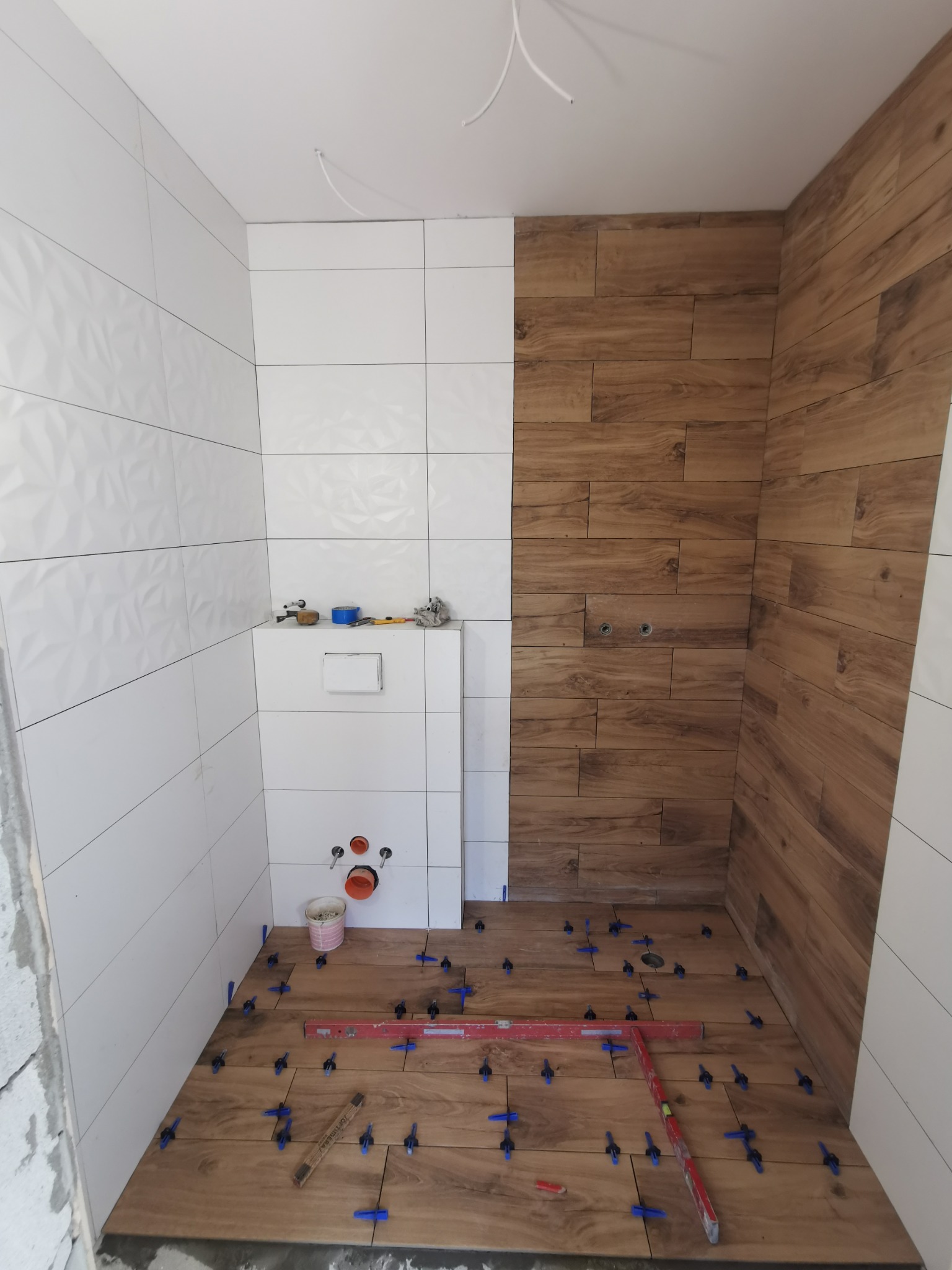 Układanie płytek imitujących drewno na podłodze i białych płytek na ścianach w łazience, widoczne poziomica, krzyżyki dystansowe i narzędzia glazurnicze.