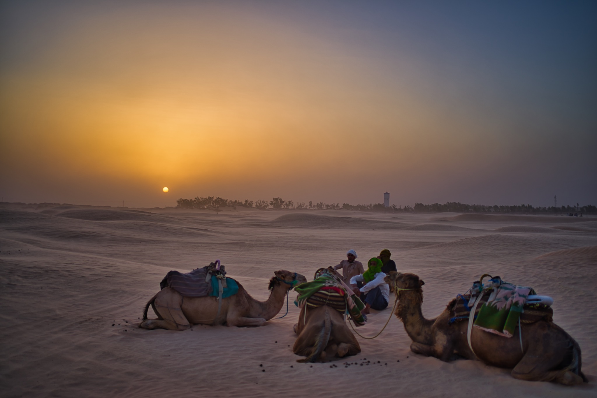 Zachód słońca nad Saharą, wielbłądy odpoczywają na piasku, w tle siedząca grupa ludzi i odległa wieża ciśnień.