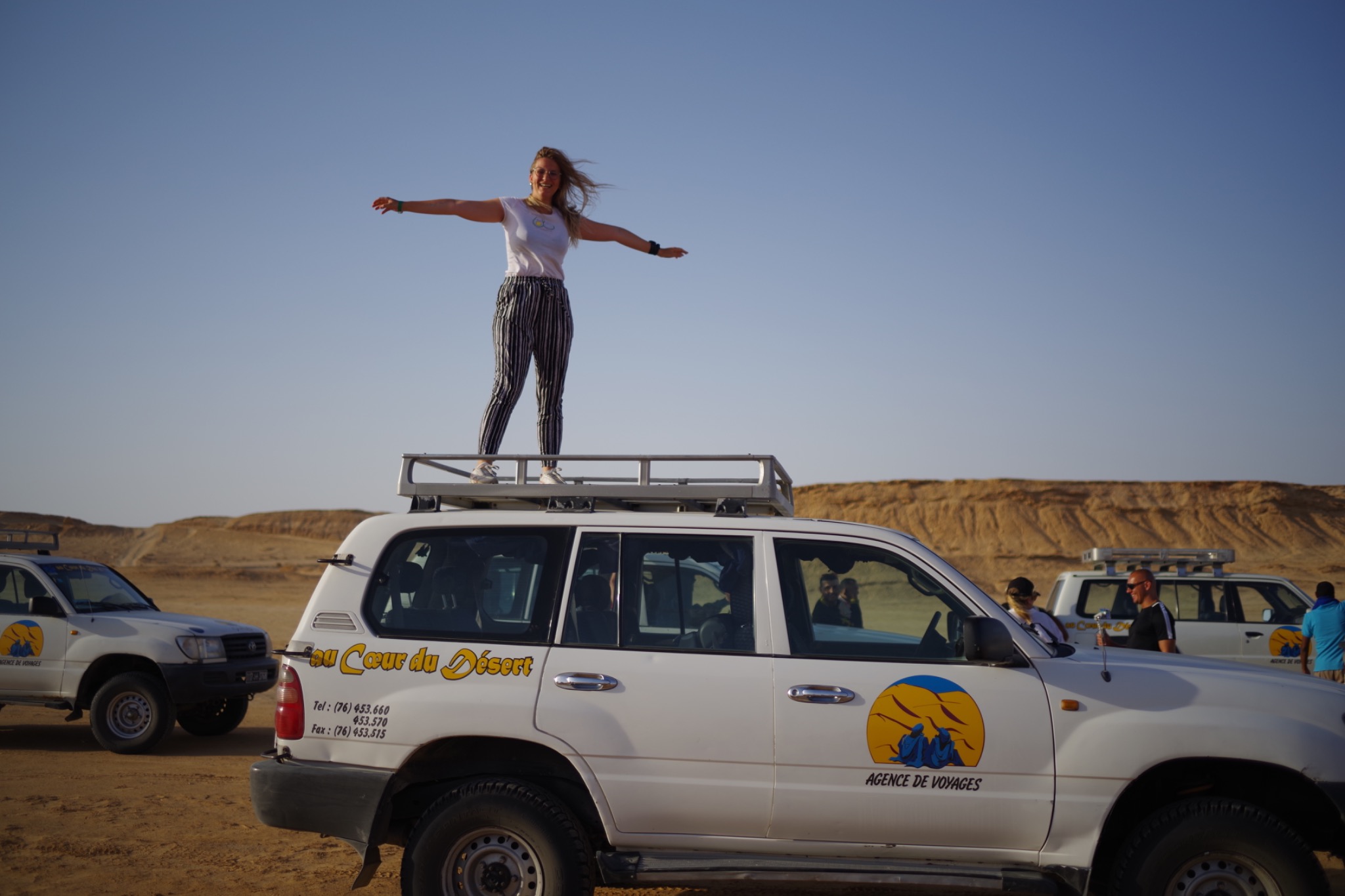 Kobieta w paski na dachu białego samochodu terenowego z logiem biura podróży na tle pustynnego krajobrazu i błękitnego nieba.