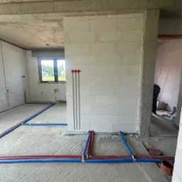 Instalacja ogrzewania podłogowego w surowym wnętrzu. Czerwono-niebieskie rury ułożone na szarym betonie, ściany z białego bloczka, widoczne pionowe bruzdy pod instalacje.