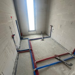 Pro-Technik Damian Skoczylas - Surowy stan łazienki z rozprowadzoną instalacją wodną (czerwone i niebieskie rury) oraz kanalizacyjną. Widoczne białe ściany z betonu komórkowego i okno.