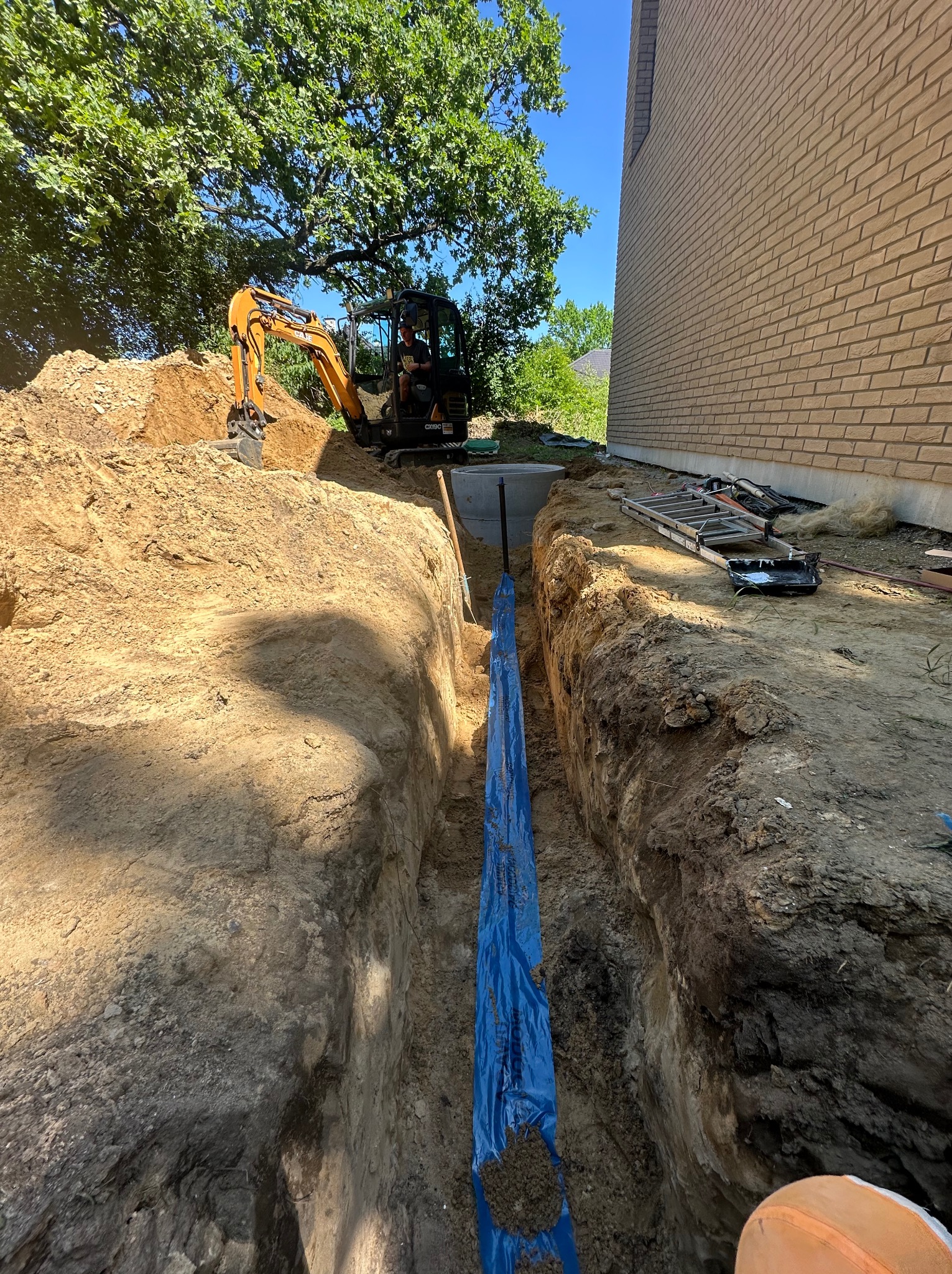 Wykop pod instalacje przy budynku z cegły, zabezpieczony folią. Na górze koparka i elementy szalunku, widoczne narzędzia. Prace ziemne w toku.