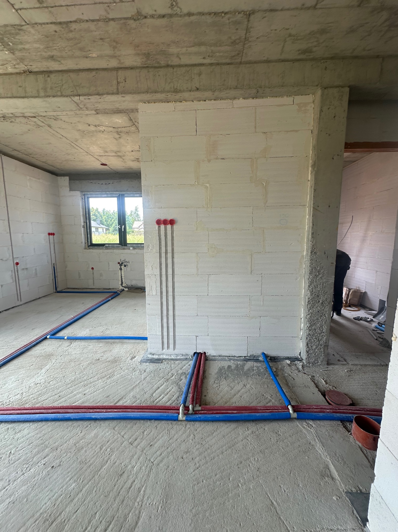 Instalacja ogrzewania podłogowego w surowym wnętrzu. Czerwono-niebieskie rury ułożone na szarym betonie, ściany z białego bloczka, widoczne pionowe bruzdy pod instalacje.