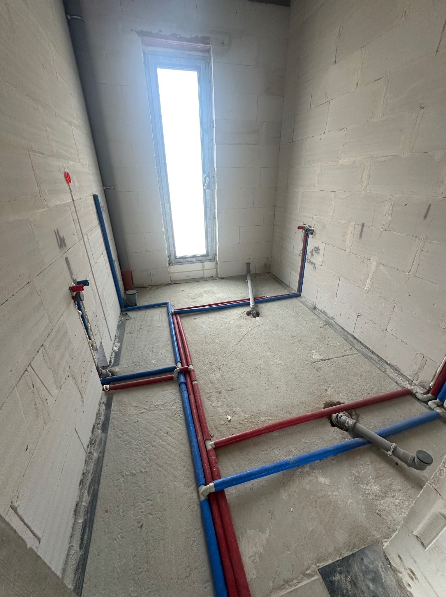 Surowy stan łazienki z rozprowadzoną instalacją wodną (czerwone i niebieskie rury) oraz kanalizacyjną. Widoczne białe ściany z betonu komórkowego i okno.