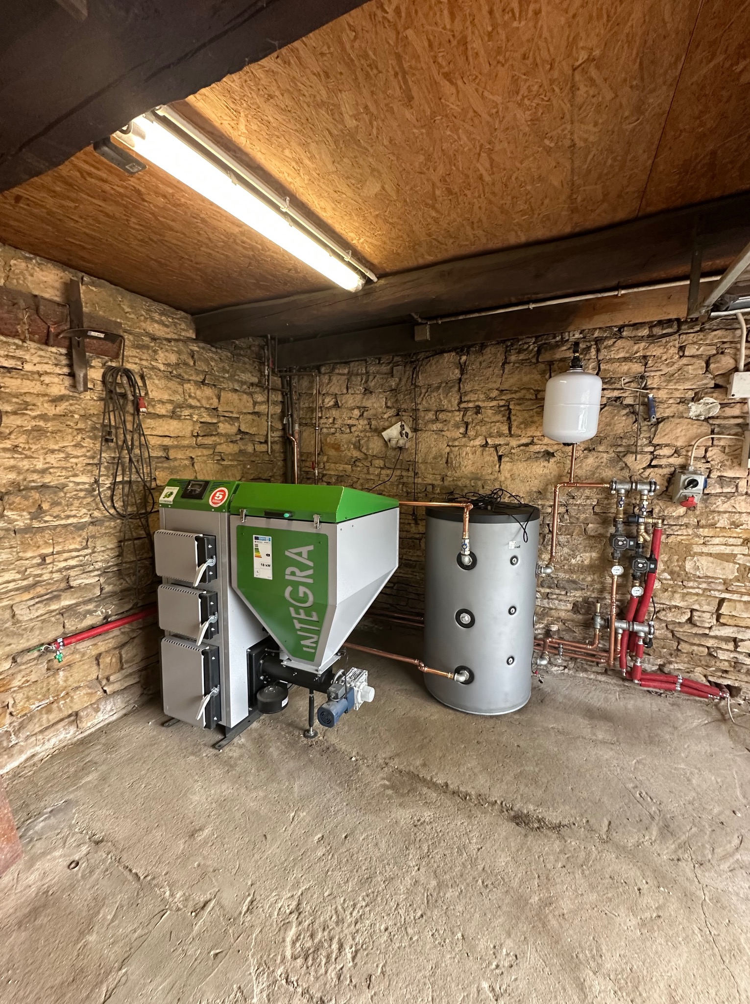 Nowoczesny piec grzewczy Integra z zbiornikiem buforowym w kotłowni o kamiennych ścianach, oświetlony jarzeniówką. Widoczne przyłącza hydrauliczne.