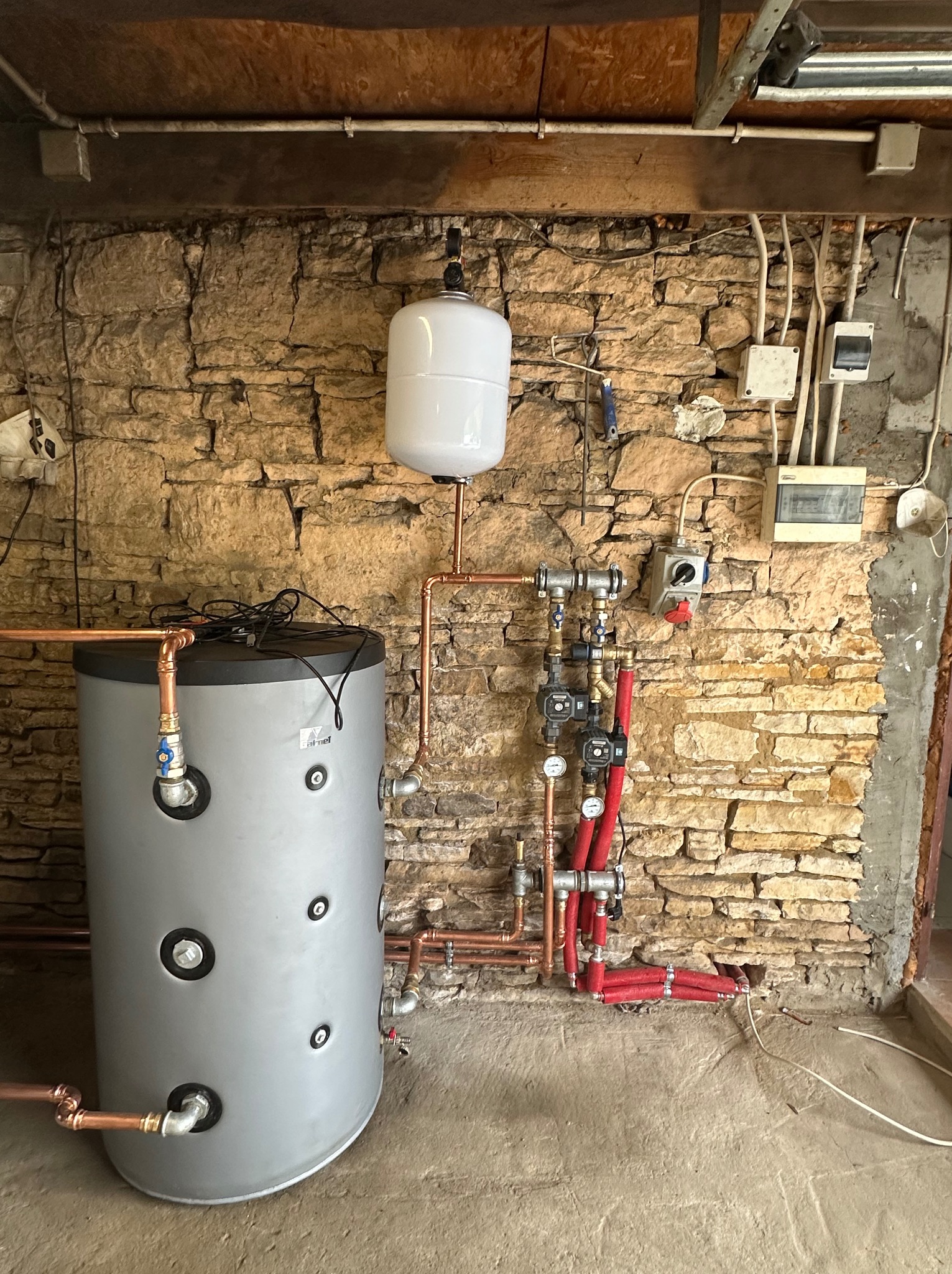 Szary bojler z osprzętem i zbiornikiem wyrównawczym na tle kamiennej ściany. Miedziane rury, izolacja termiczna. Instalacja w piwnicy, widoczne przewody elektryczne.