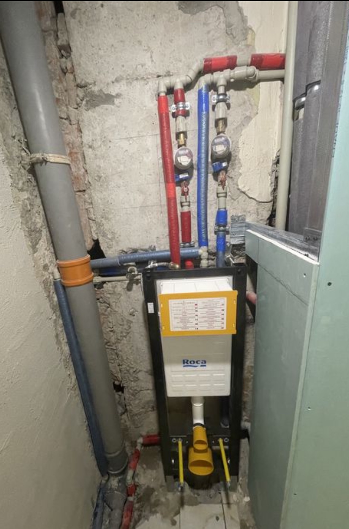 Instalacja podtynkowa WC marki Roca z widocznymi rurami doprowadzającymi wodę (czerwona i niebieska) w surowym wnętrzu, przed zabudową płytami.