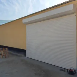 Zamknięta biała roleta zewnętrzna na oknie budynku z żółtą elewacją, widoczne drewniane kozły i zwinięty przedłużacz elektryczny na betonowym podłożu.