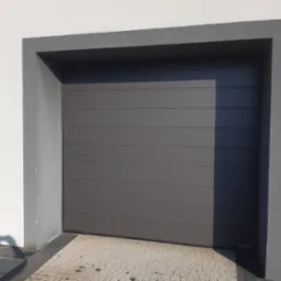 Zamknięta, segmentowa brama garażowa w kolorze grafitowym, z poziomymi przetłoczeniami, osadzona w szarej ramie na tle białej ściany budynku. W rogu widoczny cień fotografa.