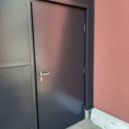 Ciemnoszara brama przemysłowa z klamką i zamkiem, zamontowana w budynku z elewacją z blachy trapezowej i fragmentem ściany otynkowanej na ceglasto-czerwono.