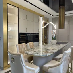 Luksusowa kuchnia z wyspą o blacie z granitu, beżowymi krzesłami barowymi z ozdobnymi pierścieniami i złotymi akcentami, w tym okapem i obramowaniem szafek.
