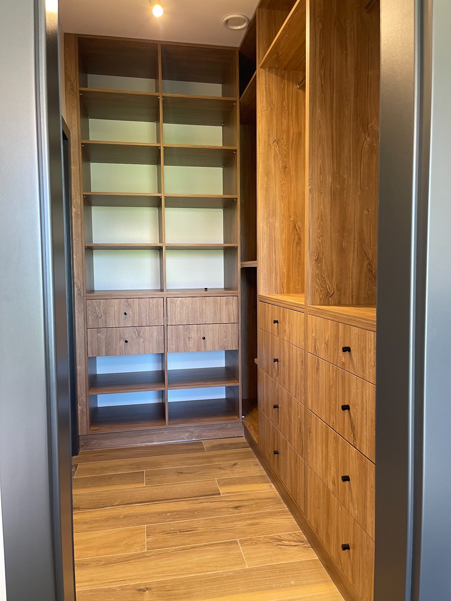 Wnętrze garderoby z zabudową z forniru w odcieniach drewna, półki i szuflady z czarnymi gałkami, oświetlenie punktowe, widok z perspektywy wejścia.