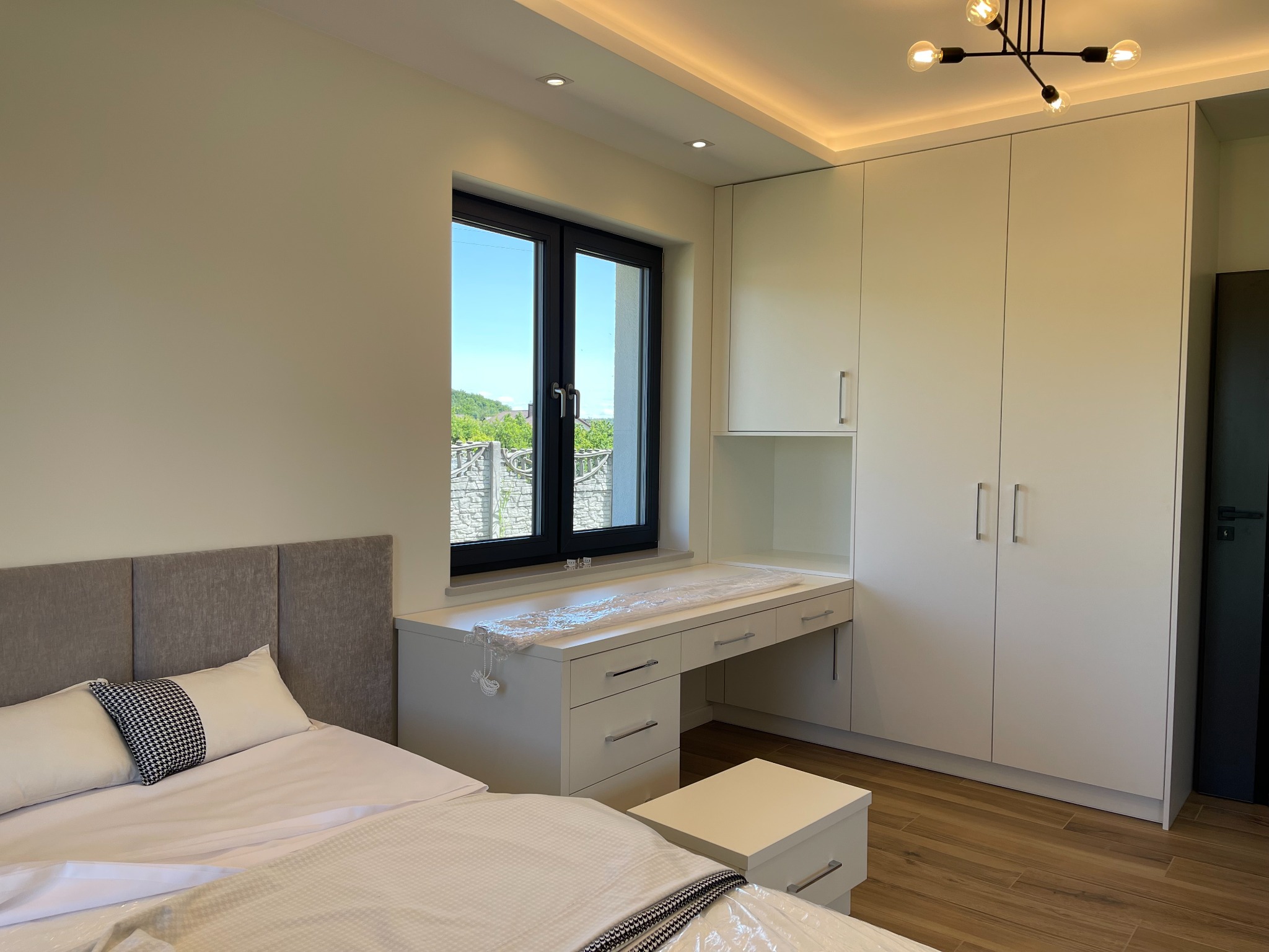 Jasna sypialnia z biurkiem pod oknem, białymi szafami i łóżkiem z szarą ramą, widok na zieleń za oknem.