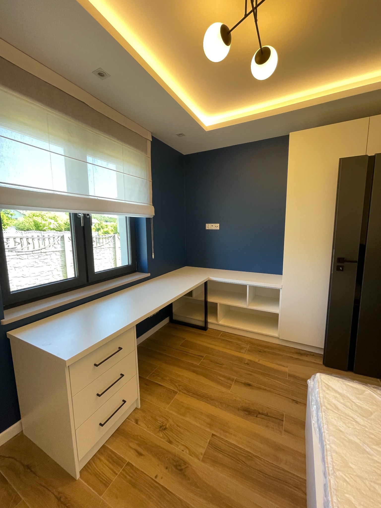 Nowoczesny pokój z biurkiem narożnym w kolorze białym, granatowa ściana, drewnopodobna podłoga i oświetlenie LED na suficie.