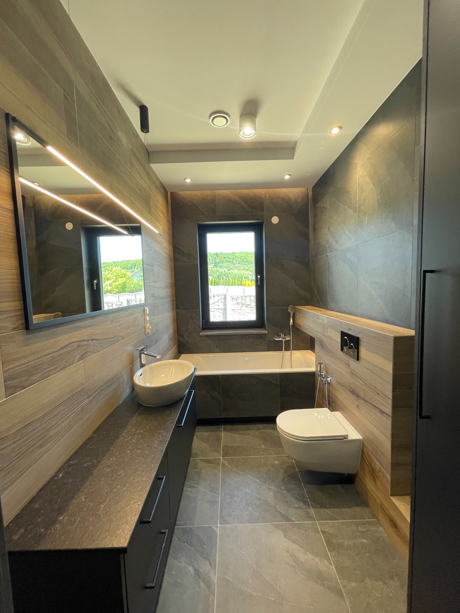 Nowoczesna łazienka z płytkami imitującymi drewno i kamień, podświetlane lustro, wisząca toaleta i umywalka nablatowa, widok na zieleń za oknem.