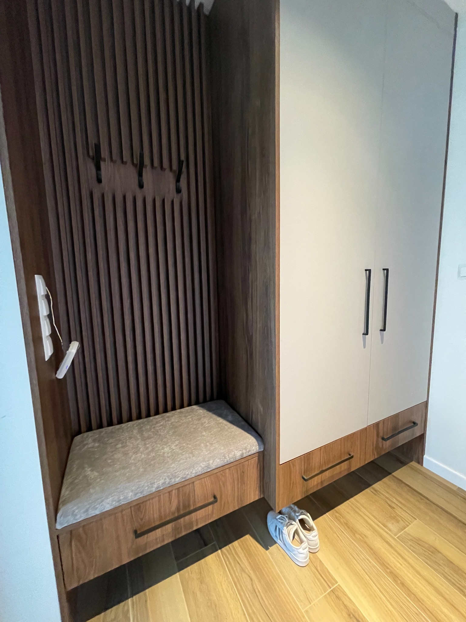 Wnęka w przedpokoju z siedziskiem, wieszakami na ubrania i szafką w kolorach drewna i bieli, uzupełniona o parę białych butów sportowych.