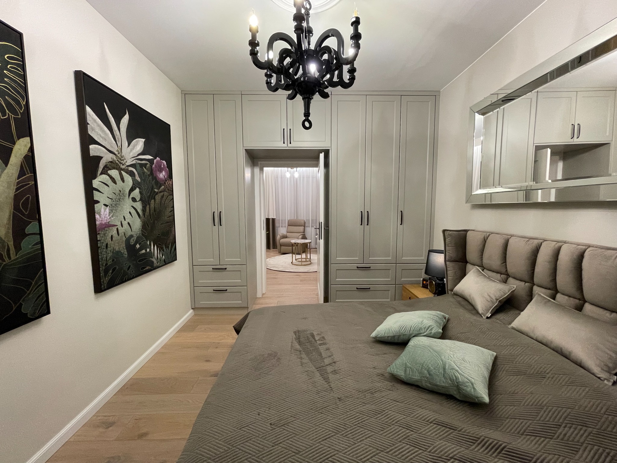 Nowoczesna sypialnia z dużym łóżkiem, szarą tapicerowaną ramą, szafą w zabudowie, czarnym żyrandolem i obrazem z motywami roślinnymi.