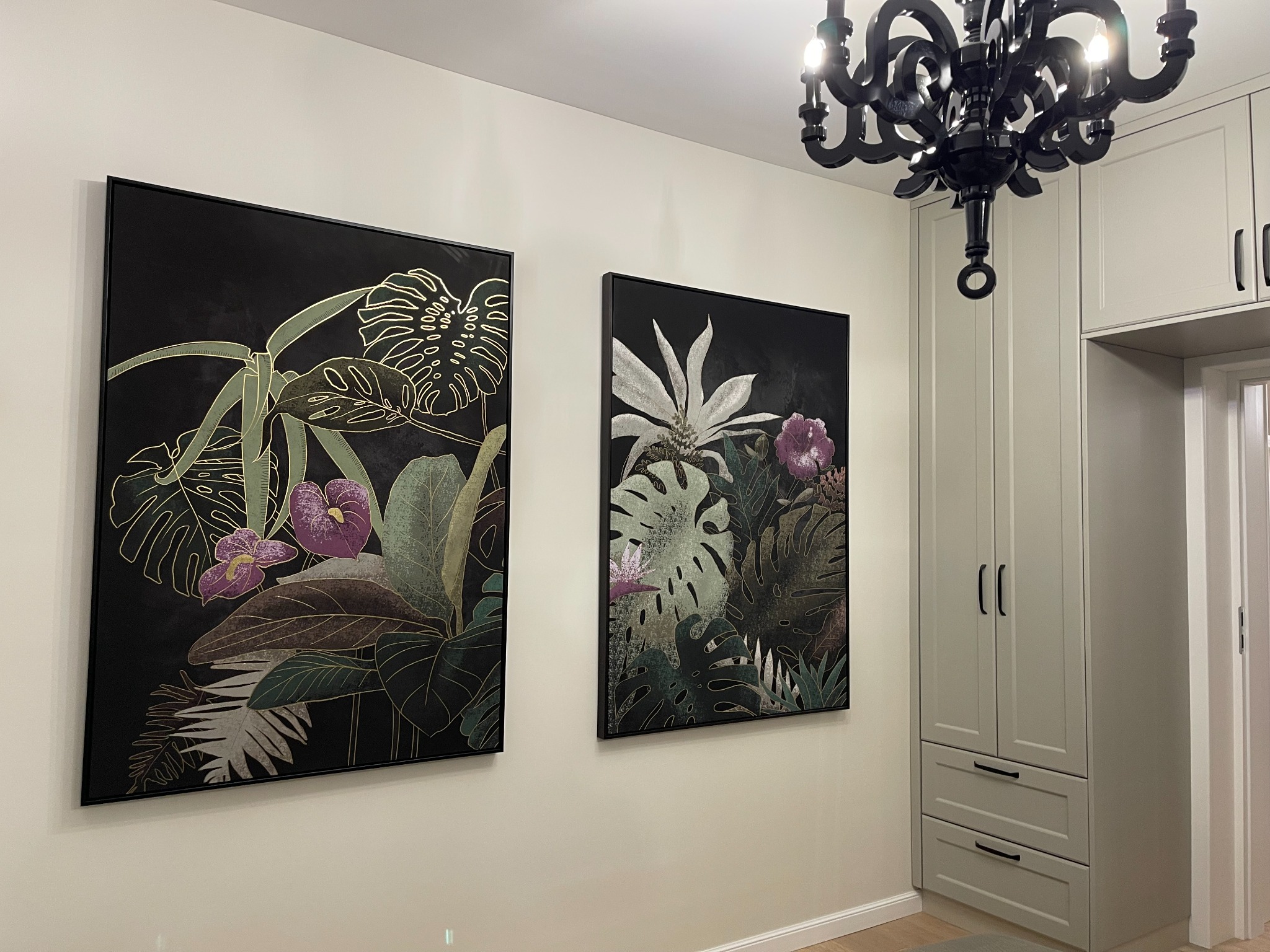 Dwa obrazy w czarnych ramach z motywem tropikalnych roślin i kwiatów na ścianie, widoczny fragment czarnego żyrandola i szara szafa w tle.