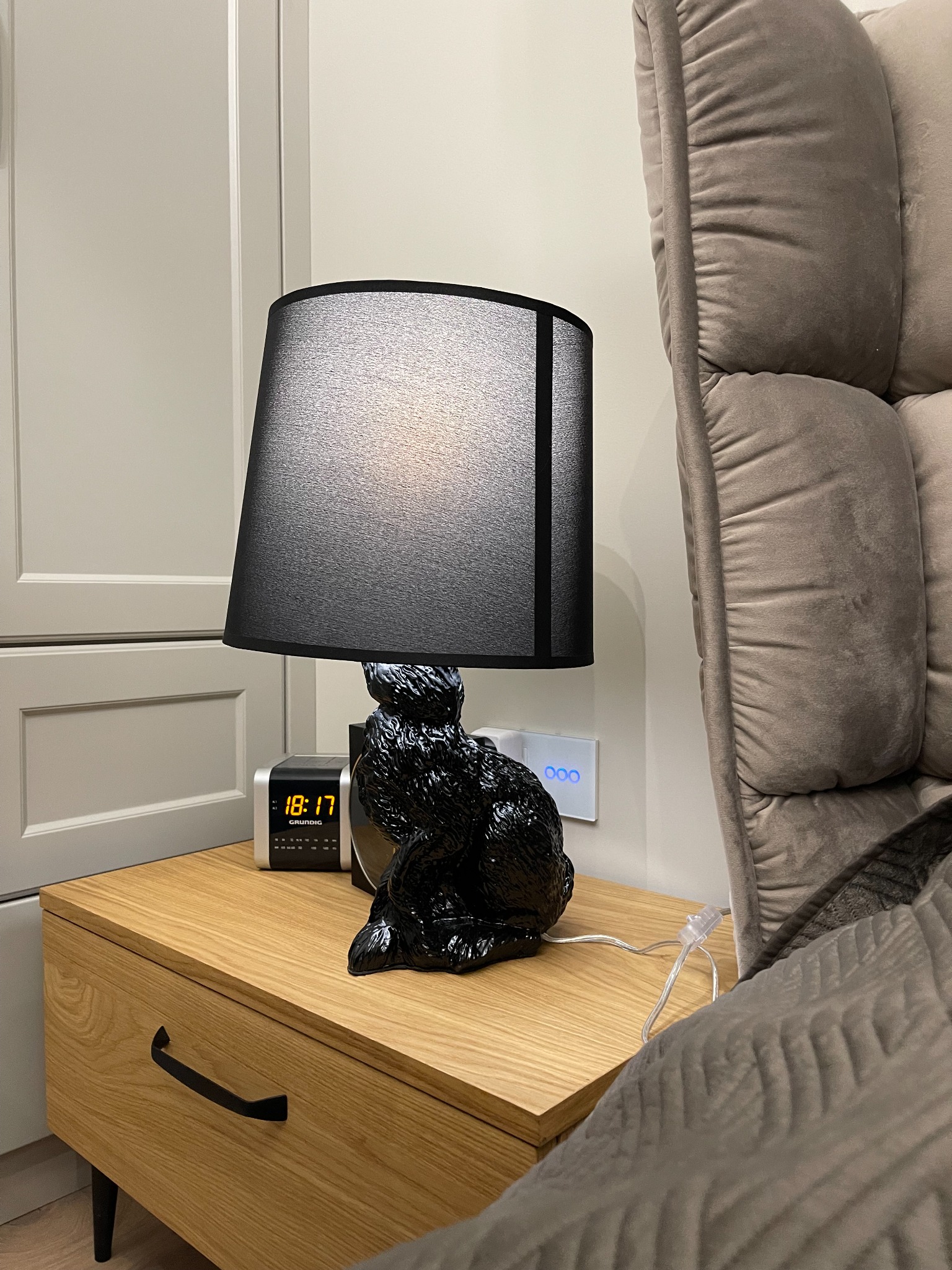 Stylowa lampka nocna w kształcie czarnego królika z abażurem, stojąca na drewnianej szafce nocnej obok cyfrowego budzika i fragmentu szarego fotela z poduszkami.