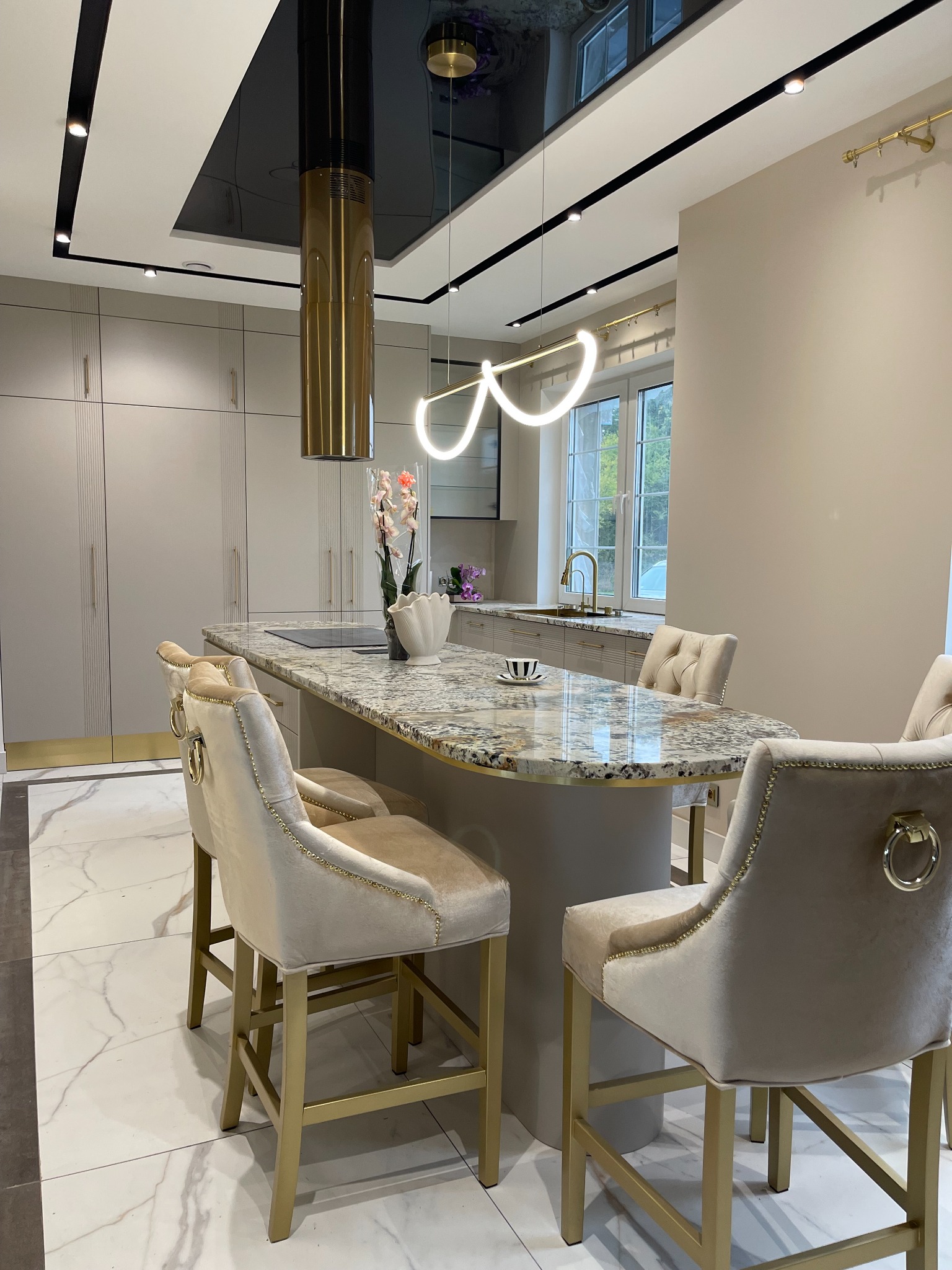 Elegancka kuchnia z wyspą z blatem z granitu, hokery z welurowym obiciem w kolorze ecru i złotymi nogami, okap w kolorze złotym, nowoczesne oświetlenie.