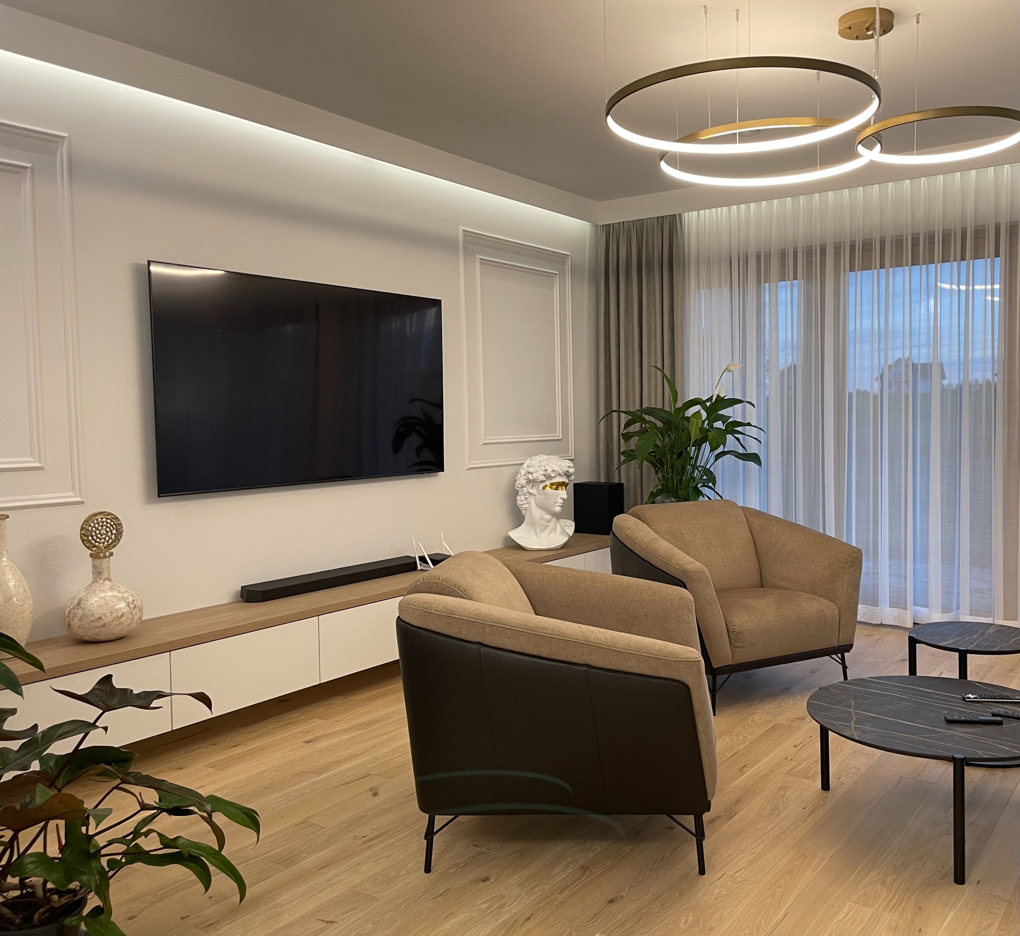 Nowoczesny salon z drewnianą podłogą, beżowymi fotelami, okrągłymi stolikami z czarnym marmurkowym blatem i dekoracyjnym oświetleniem w postaci trzech pierścieni.