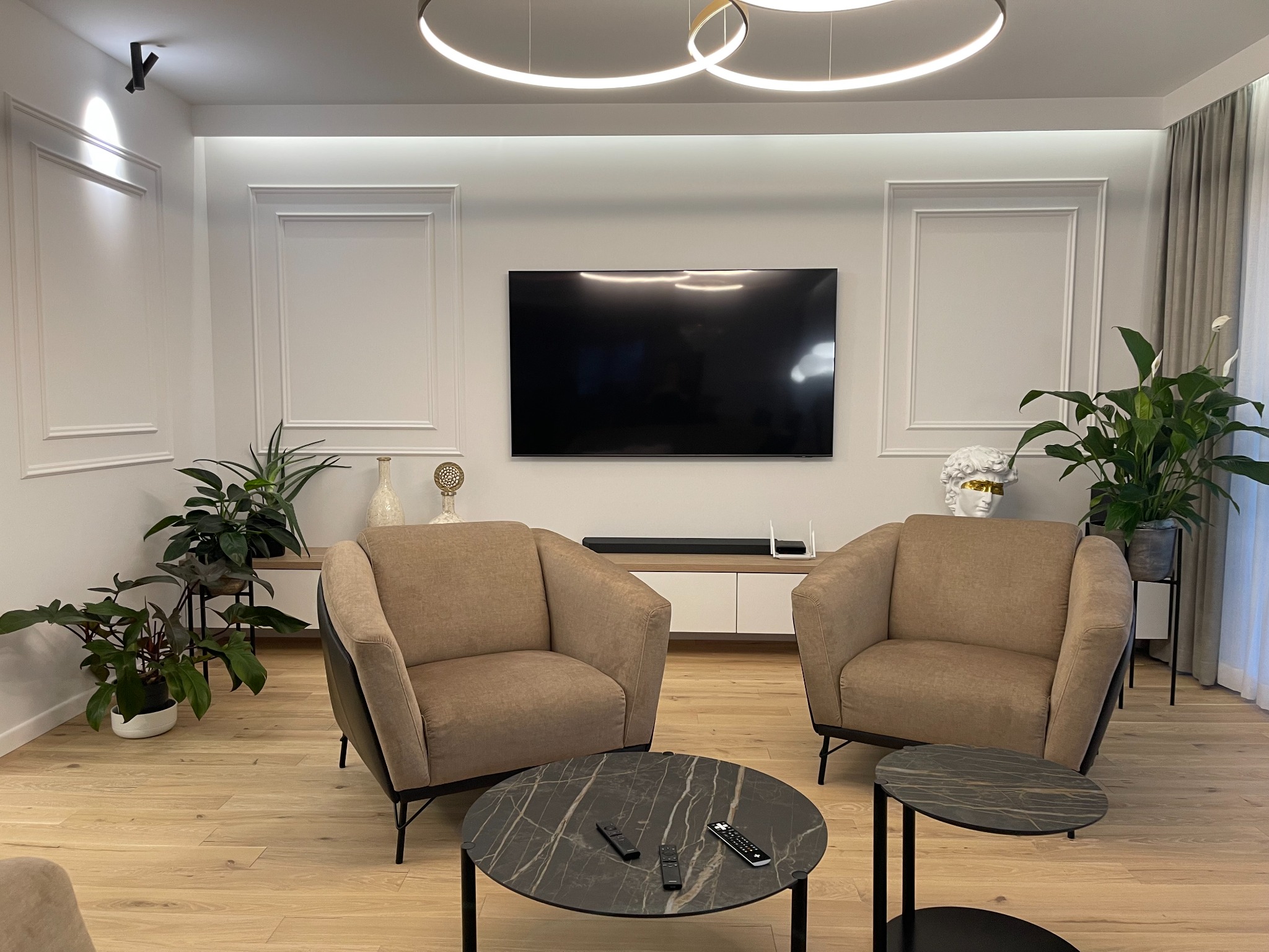 Jasny salon z parkietem, dwoma fotelami, okrągłymi stolikami z marmurowym wzorem, telewizorem na ścianie, dekoracjami i roślinami doniczkowymi.