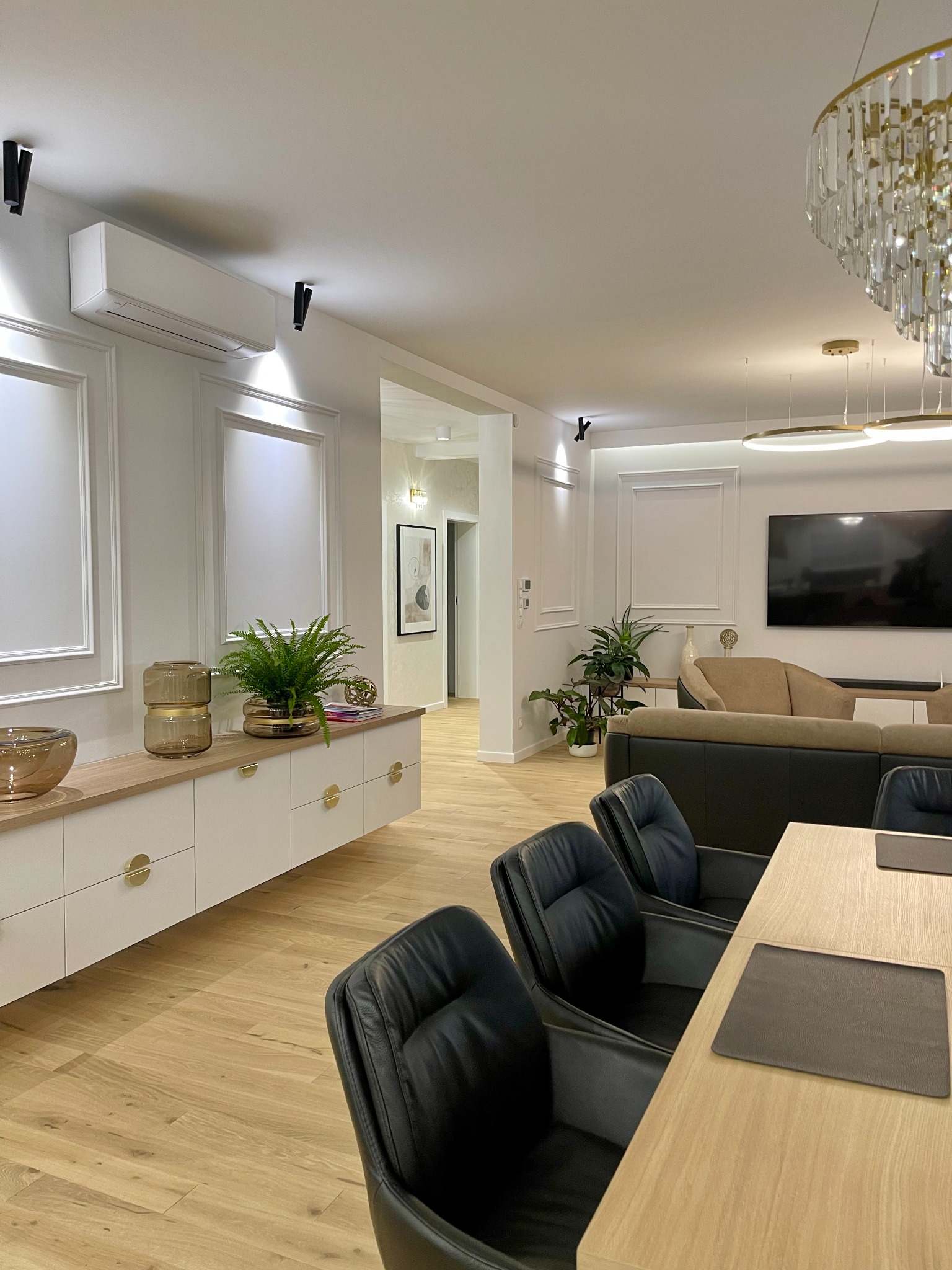 Nowoczesne wnętrze salonu z jadalnią: białe ściany z dekoracyjnymi panelami, drewniana podłoga, meble w jasnych kolorach z akcentami złota, czarne skórzane krzesła przy drewnianym stole...