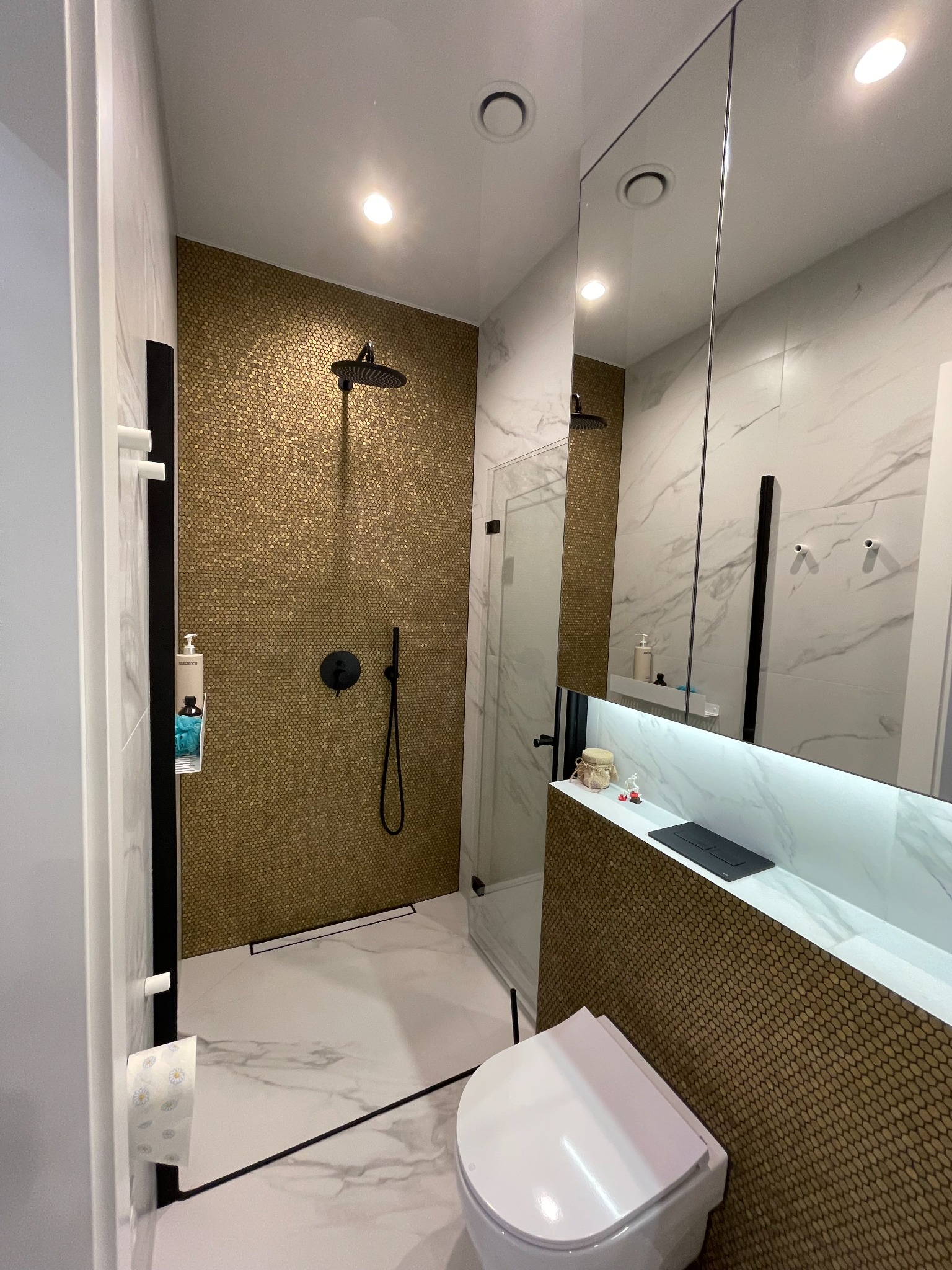 Nowoczesna łazienka z kabiną prysznicową wykończoną mozaiką w odcieniach złota, białe marmurowe płytki na ścianach, podwieszana toaleta i czarna armatura.