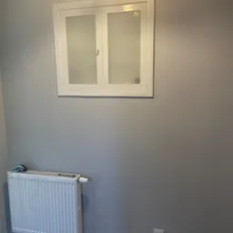 Szare wnętrze po remoncie, z białym oknem z szybami mlecznymi, białym grzejnikiem i gładzią na ścianach, widoczna listwa przypodłogowa i gniazdko elektryczne.