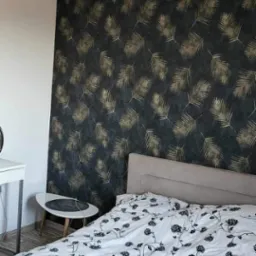 Sypialnia z tapetą w ciemne liście palmowe, białą pościelą w czarne kwiaty, stolikiem pomocniczym i lampą wiszącą w stylu industrialnym.