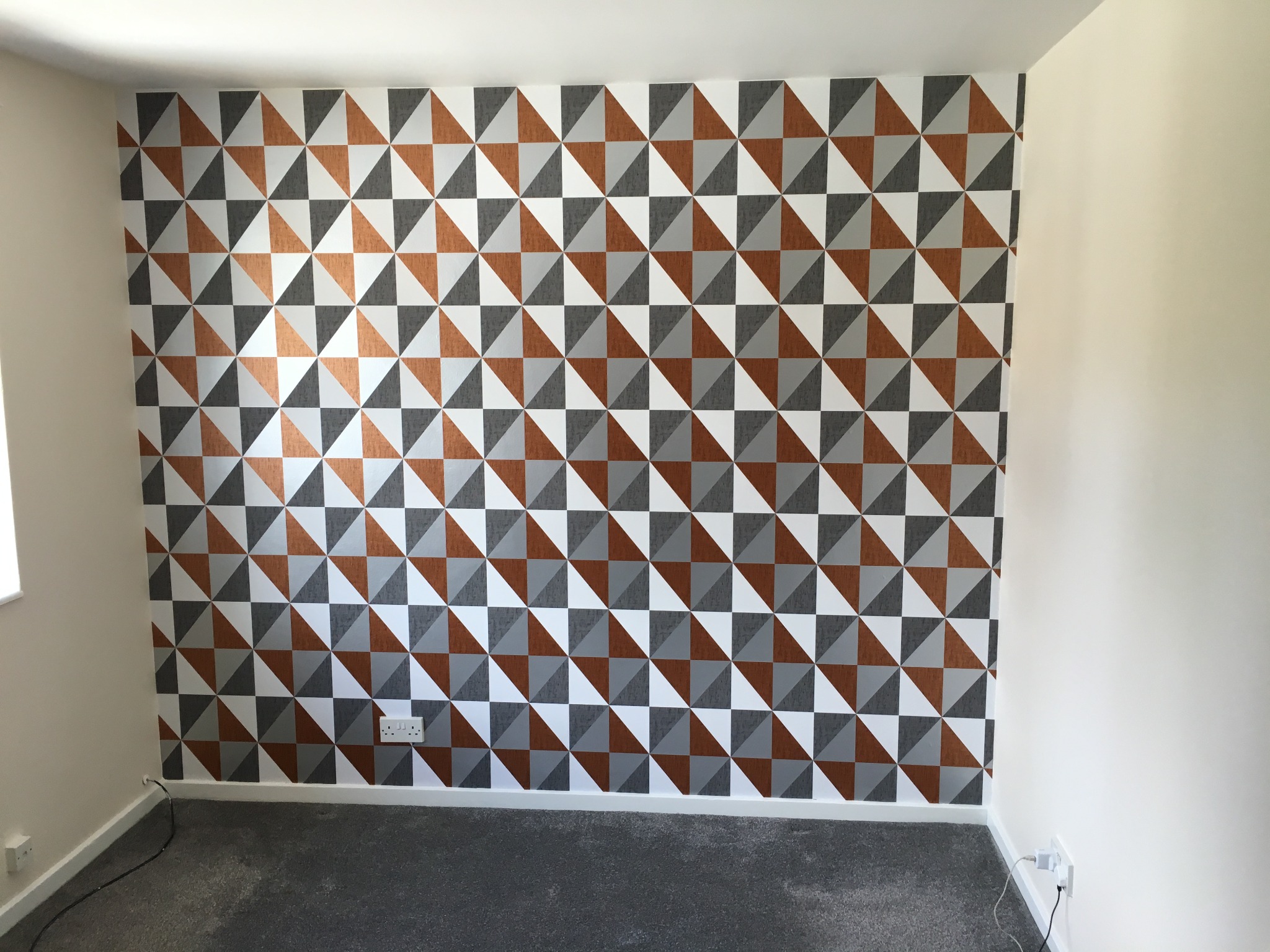 Ściana w pokoju pokryta tapetą z geometrycznym wzorem trójkątów w kolorach białym, szarym i miedzianym, szara wykładzina na podłodze, białe ściany boczne, widoczne gniazdka elektryczne.