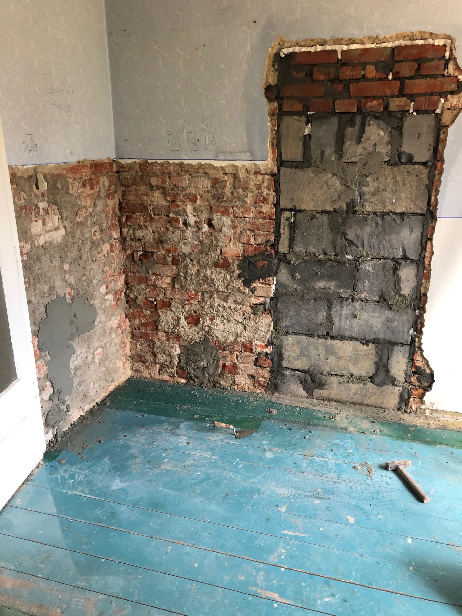 Pokój w trakcie demontażu ściany: odsłonięta cegła, fragmenty starej tapety, turkusowa podłoga z deskami, leżące narzędzia murarskie.