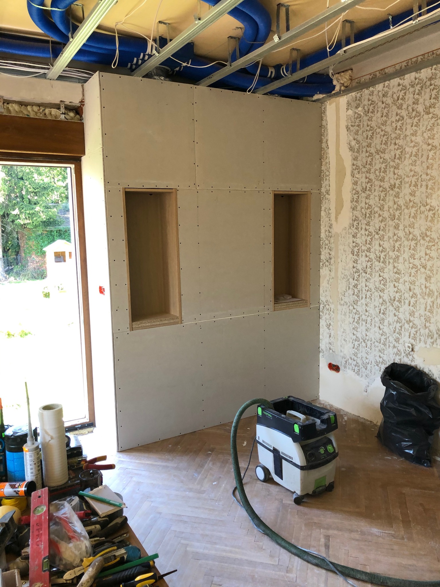 Ściana z płyt gipsowo-kartonowych w trakcie montażu, z wnękami, widoczne niebieskie rury wentylacyjne pod sufitem, resztki starej tapety na ścianie obok, podłoga parkietowa, narzędzia i odkurzacz...