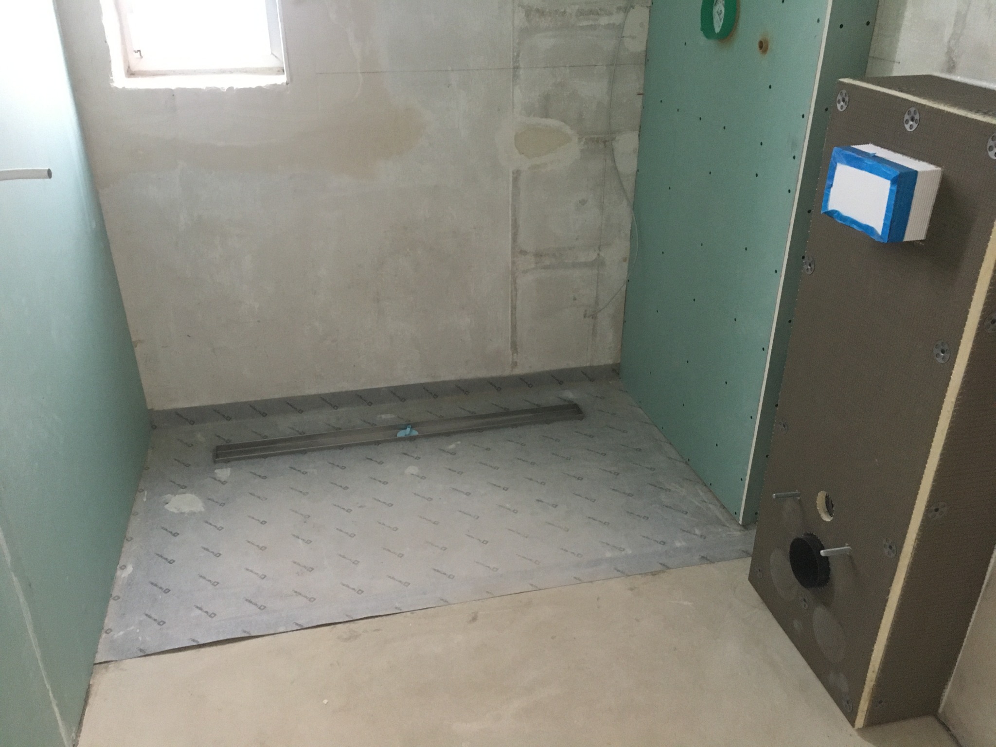 Przygotowanie do wykończenia łazienki: widok na podłogę z izolacją przeciwwodną, zamontowany odpływ liniowy oraz płyty karton-gips na ścianach, z widocznym stelażem podtynkowym WC.