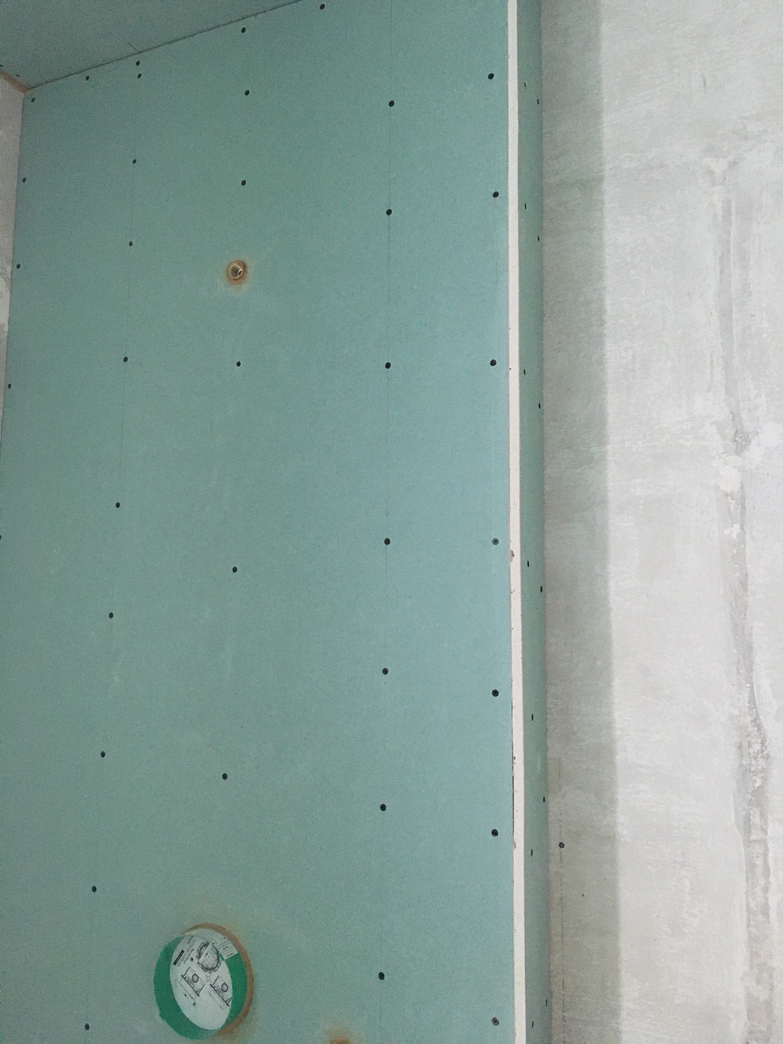 Montaż zielonej płyty gipsowo-kartonowej na ścianie, widoczne otwory po wkrętach, zabezpieczone miejsce pod instalację elektryczną, surowy stan wykończenia.