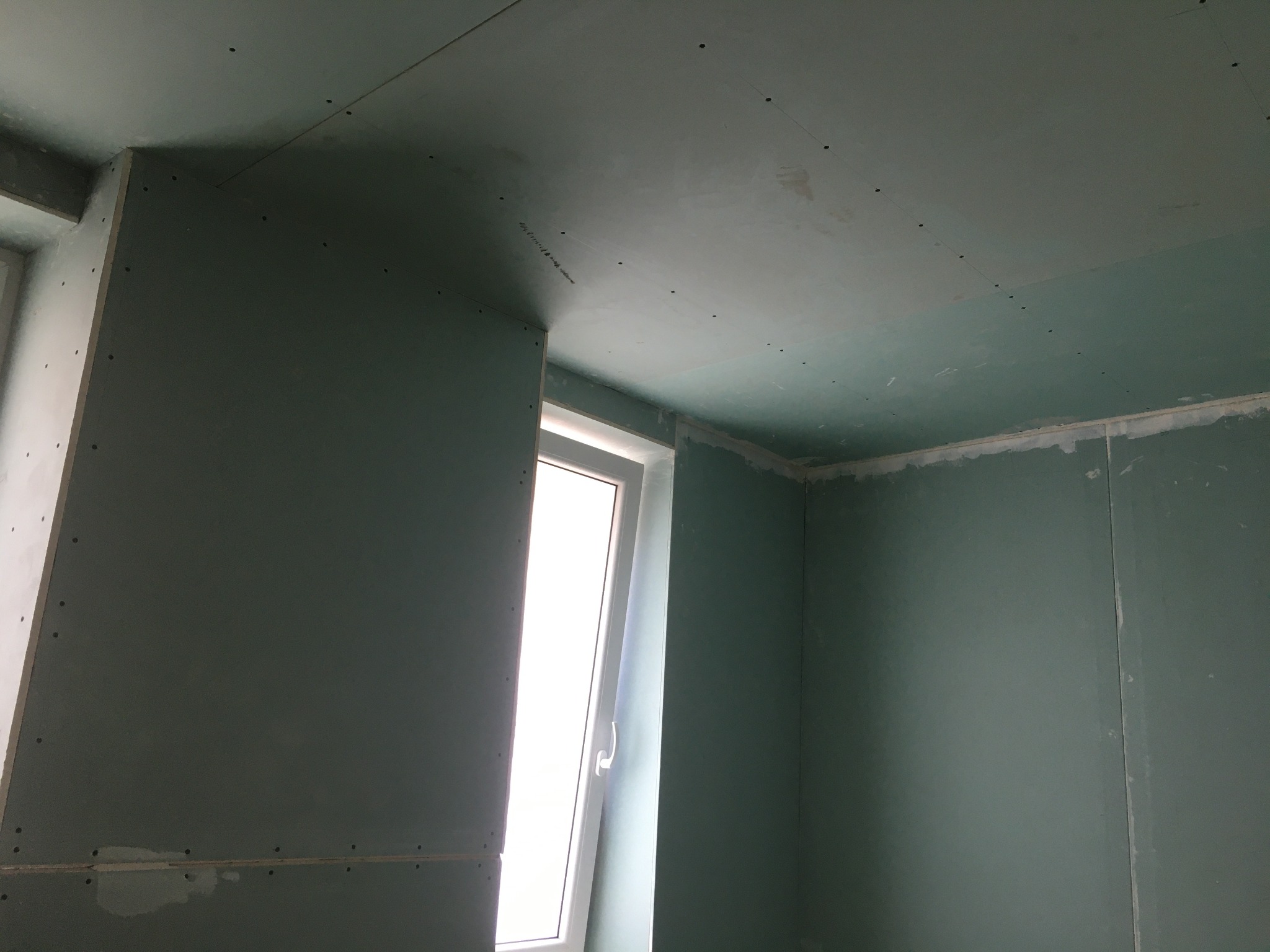 Pokój w trakcie remontu: ściany i sufit pokryte płytami gipsowo-kartonowymi, widoczne łączenia płyt i otwory po wkrętach, okno z otwartym skrzydłem wpuszcza jasne światło.