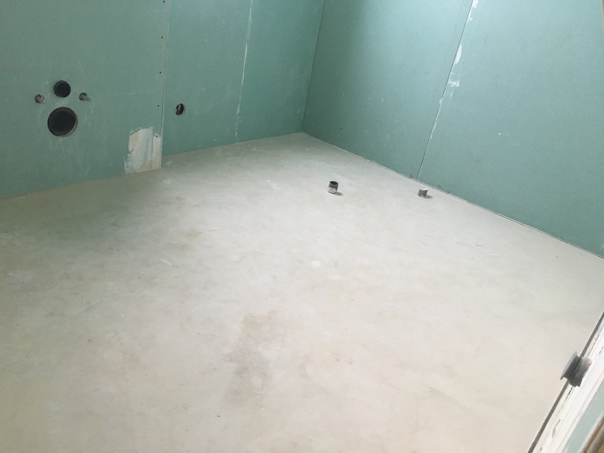 Surowe wnętrze łazienki w trakcie prac wykończeniowych, z zielonymi płytami gipsowo-kartonowymi na ścianach i wylewką podłogową, widoczne otwory pod instalacje i dwa metalowe elementy na podłodze.
