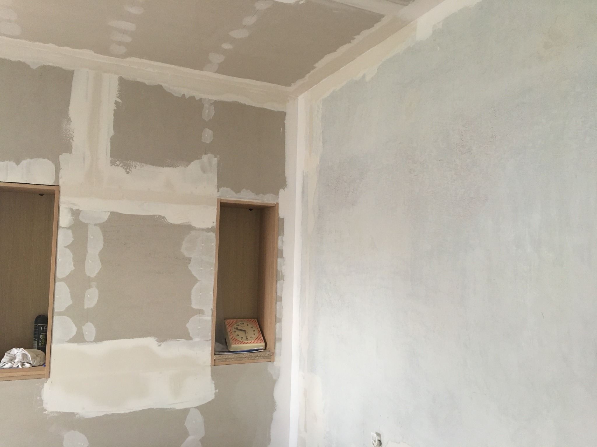 Ściana w trakcie prac wykończeniowych z widocznymi łatami gipsowymi, narożnikami i wnękami, jedna z wnęk zawiera stary zegar.