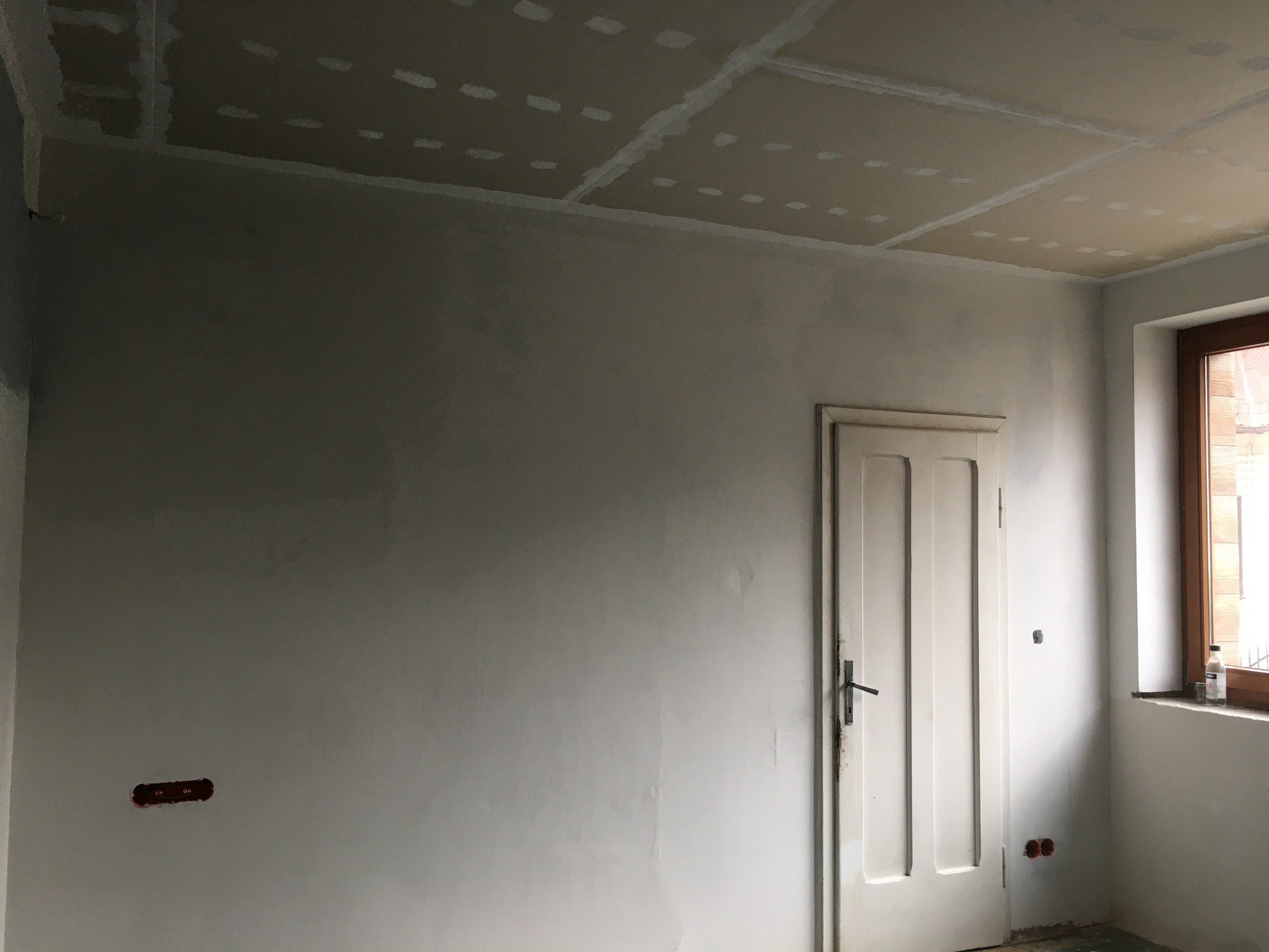 Pokój w trakcie remontu: surowa ściana po gładziowaniu, widoczne puszki elektryczne, sufit z płyt gipsowo-kartonowych ze szpachlowanymi łączeniami, stare drzwi i okno.