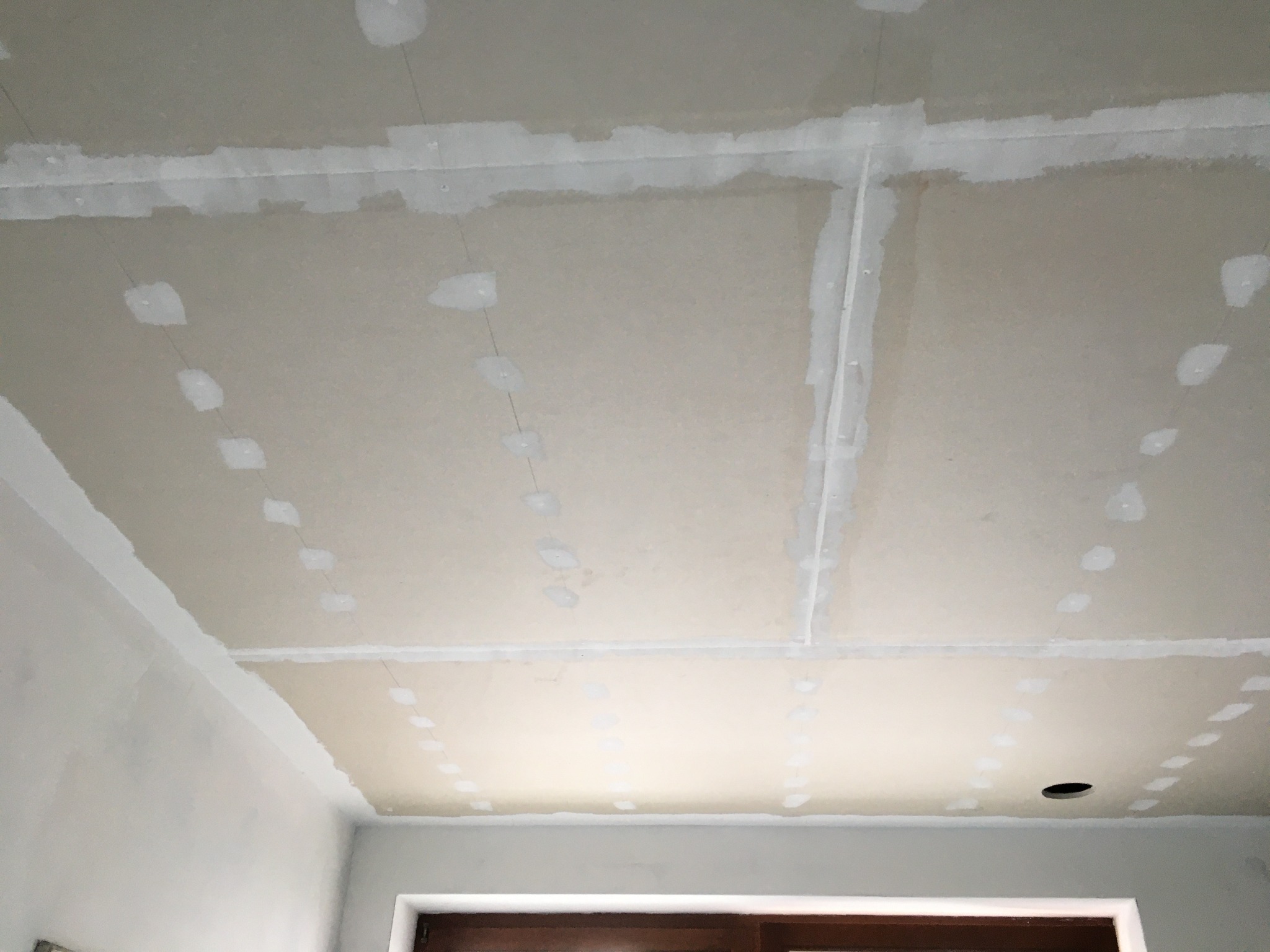 Sufit z płyt gipsowo-kartonowych w trakcie szpachlowania łączeń, widoczne ślady po wkrętach i białe pasy wzmacniające połączenia płyt, otwór na oświetlenie punktowe.