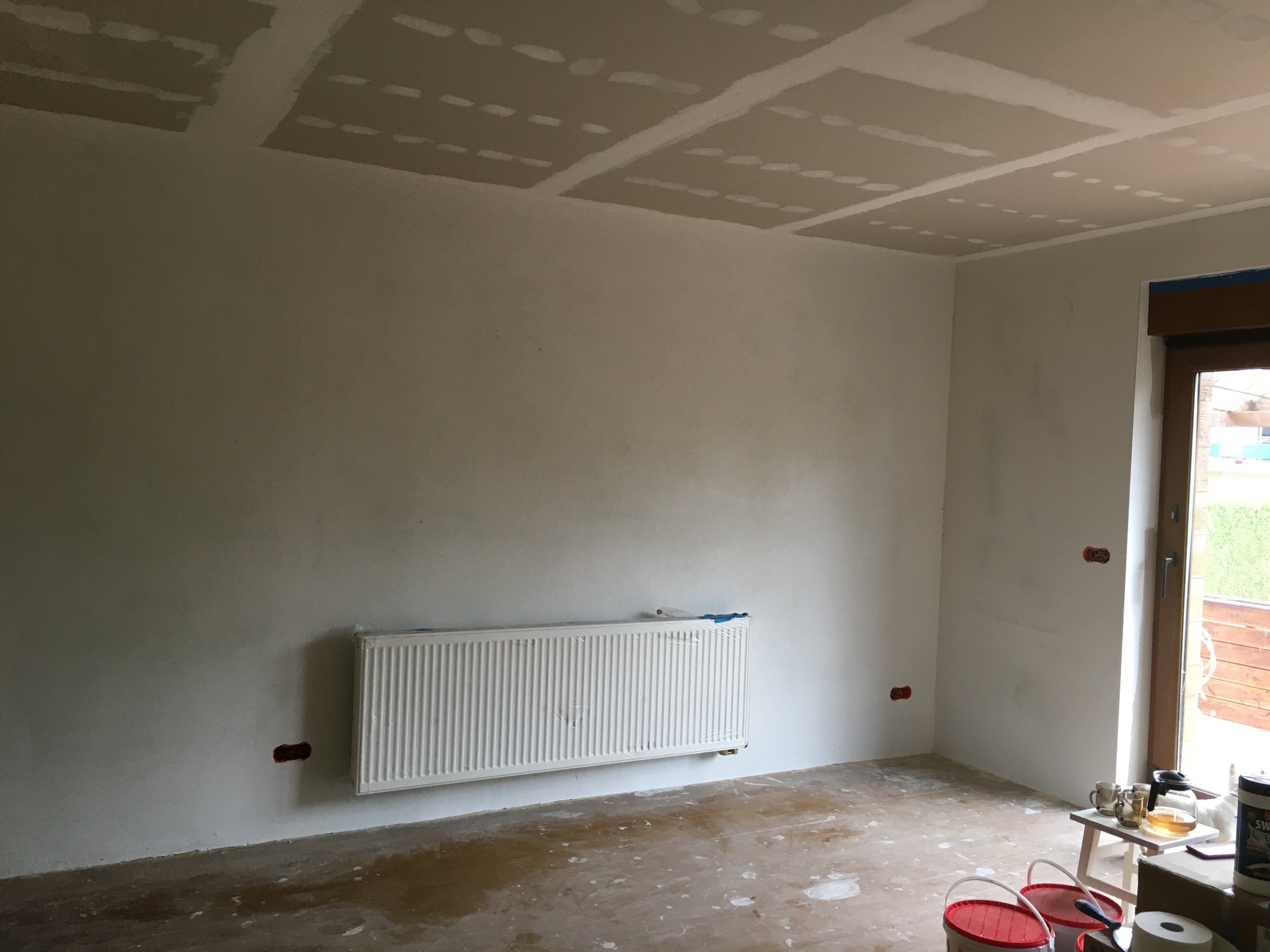 Pokój w trakcie prac wykończeniowych: ściany po gładzi, sufit z płyt kartonowo-gipsowych z widocznymi łączeniami, grzejnik owinięty folią ochronną, podłoga zabezpieczona przed zabrudzeniem...