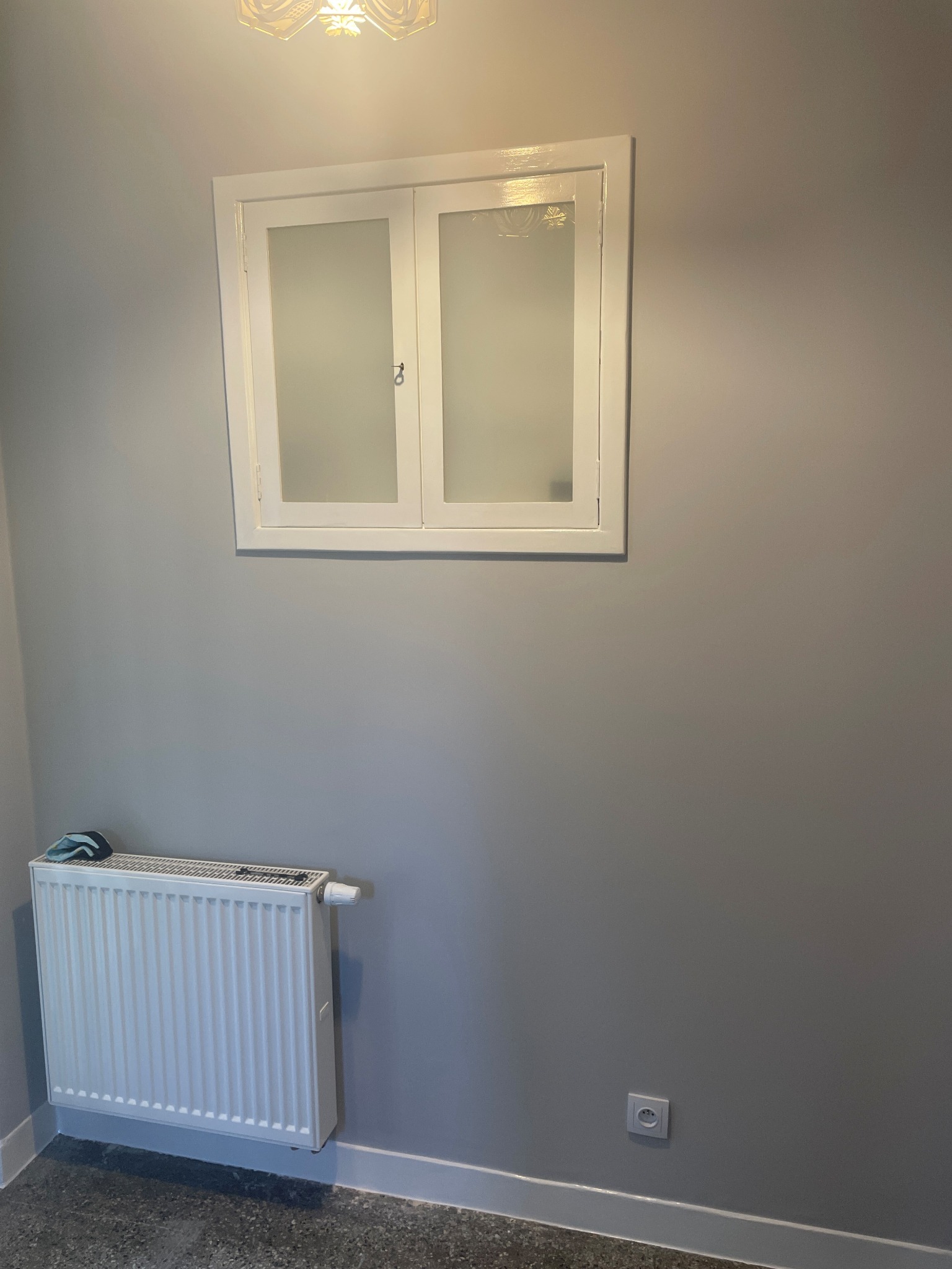 Szare wnętrze po remoncie, z białym oknem z szybami mlecznymi, białym grzejnikiem i gładzią na ścianach, widoczna listwa przypodłogowa i gniazdko elektryczne.